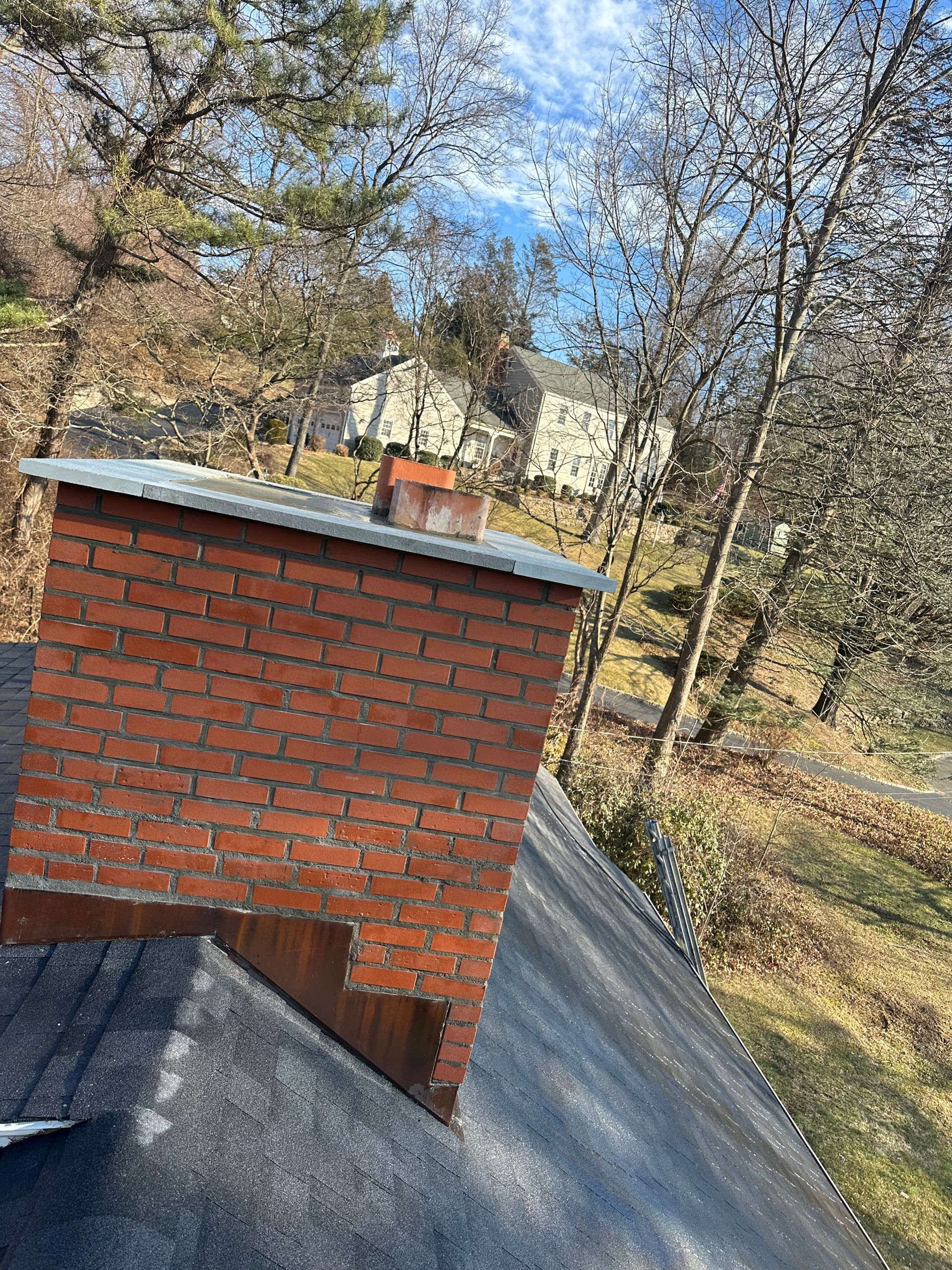 A red brick chimney with a metal cap, situated on a dark grey shingled roof with trees and a distant house in the back.