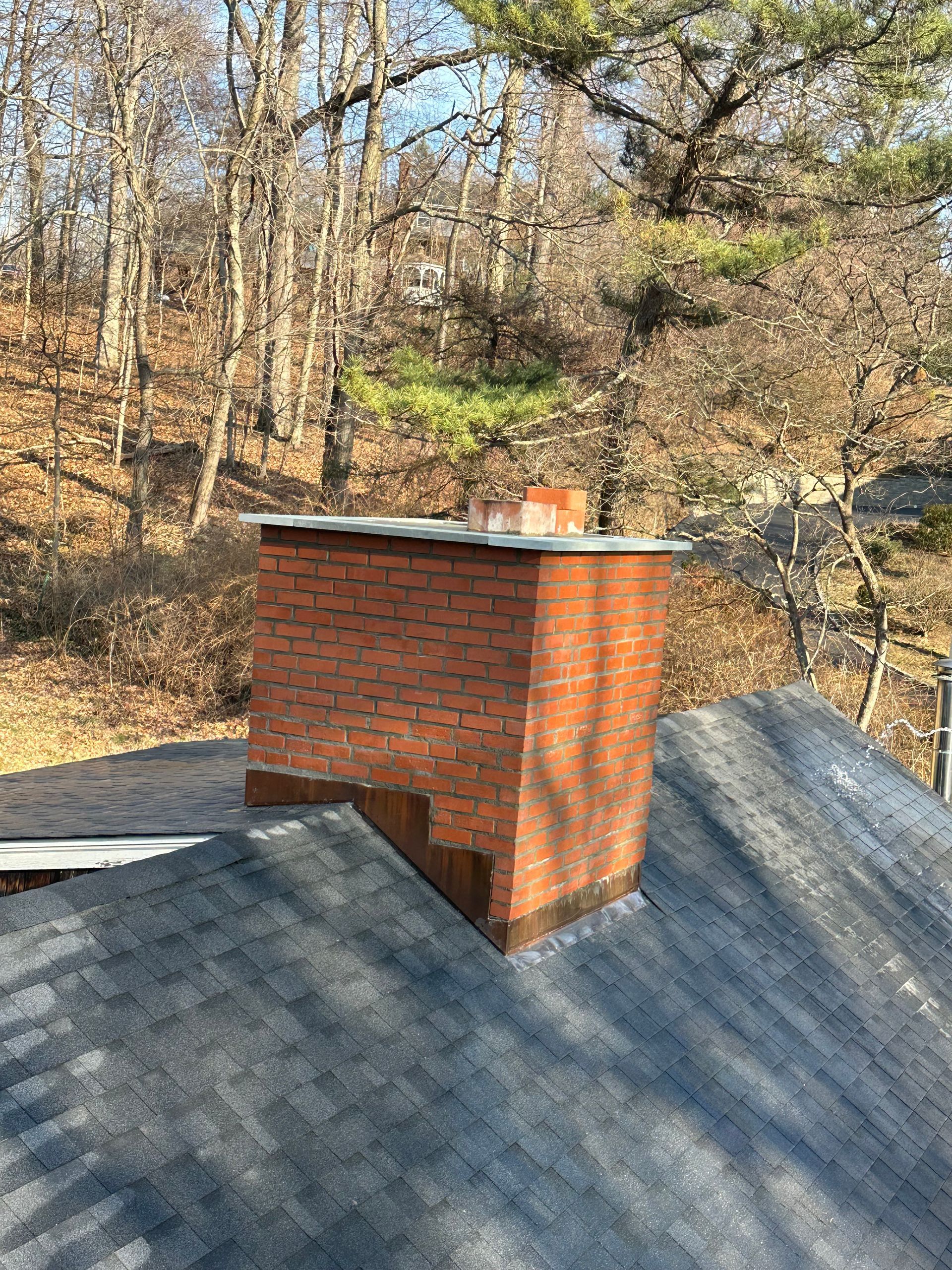 A red brick chimney with a gray metal cap rising from a dark shingled roof, surrounded by a forest of bare trees.