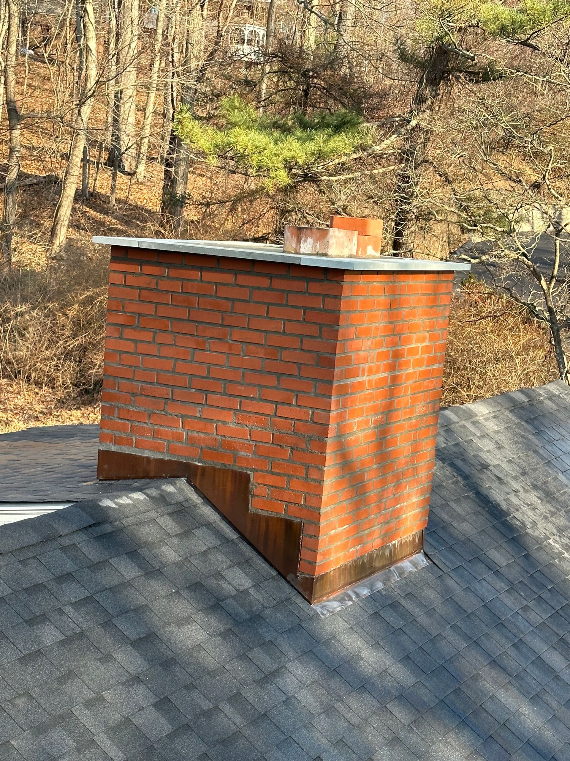 A red brick chimney with a metal cap sits on an asphalt shingle roof against a background of trees.