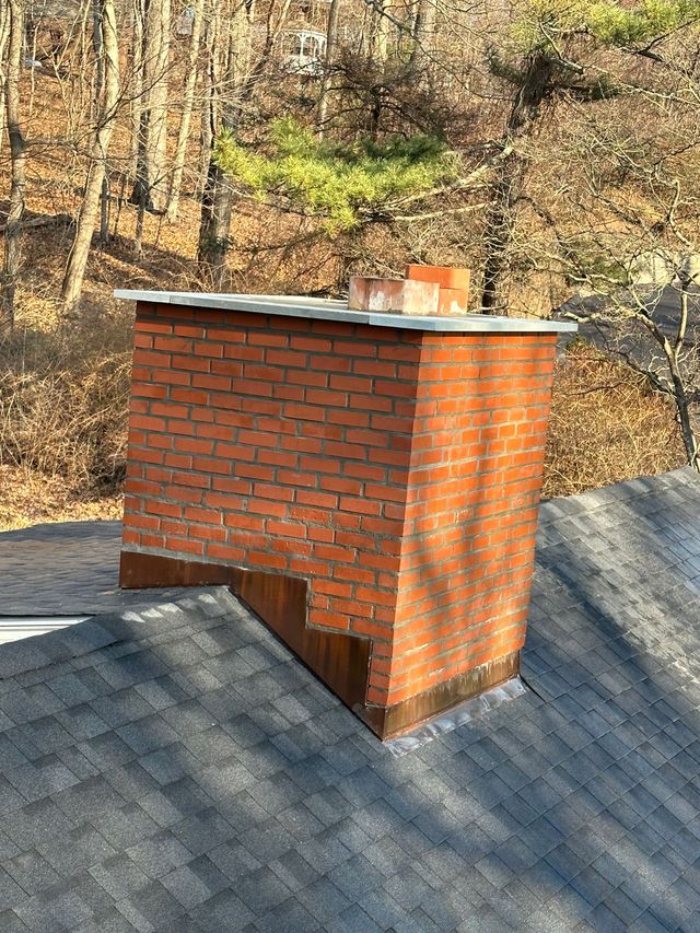 A red brick chimney with metal flashing on a gray shingled roof, set against a backdrop of trees in late autumn.