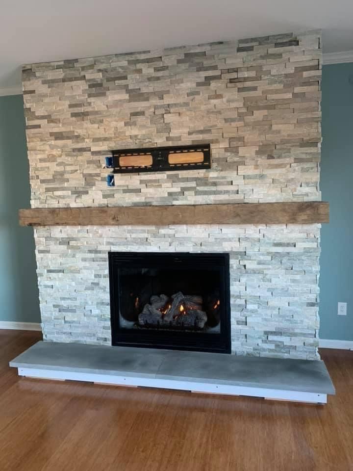 A fireplace featuring a stacked stone surround, a rustic wood mantel, a black insert, and a flat gray hearth.