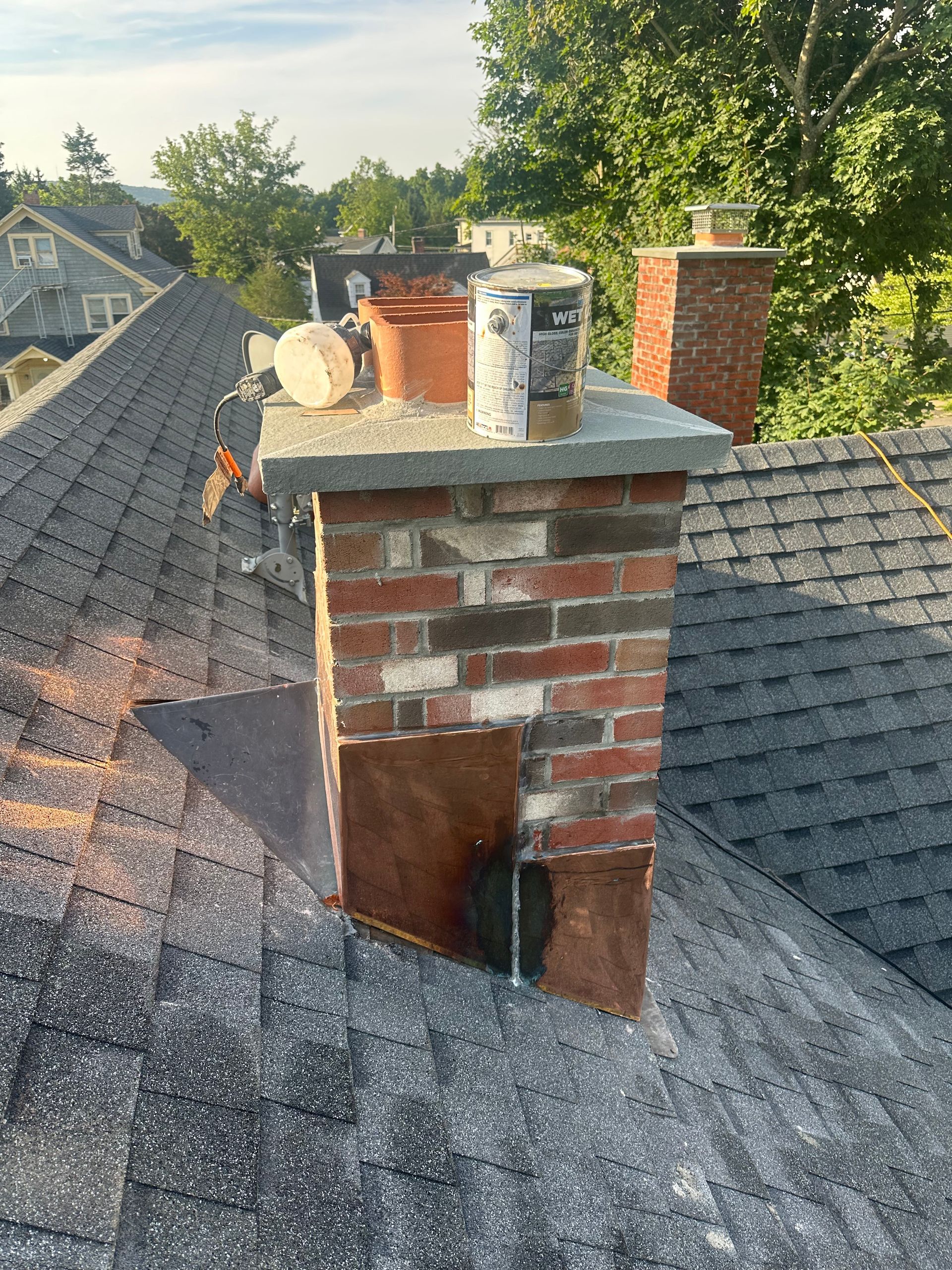 A brick chimney on a shingled roof with copper flashing, construction materials, and a paint can on the stone cap.