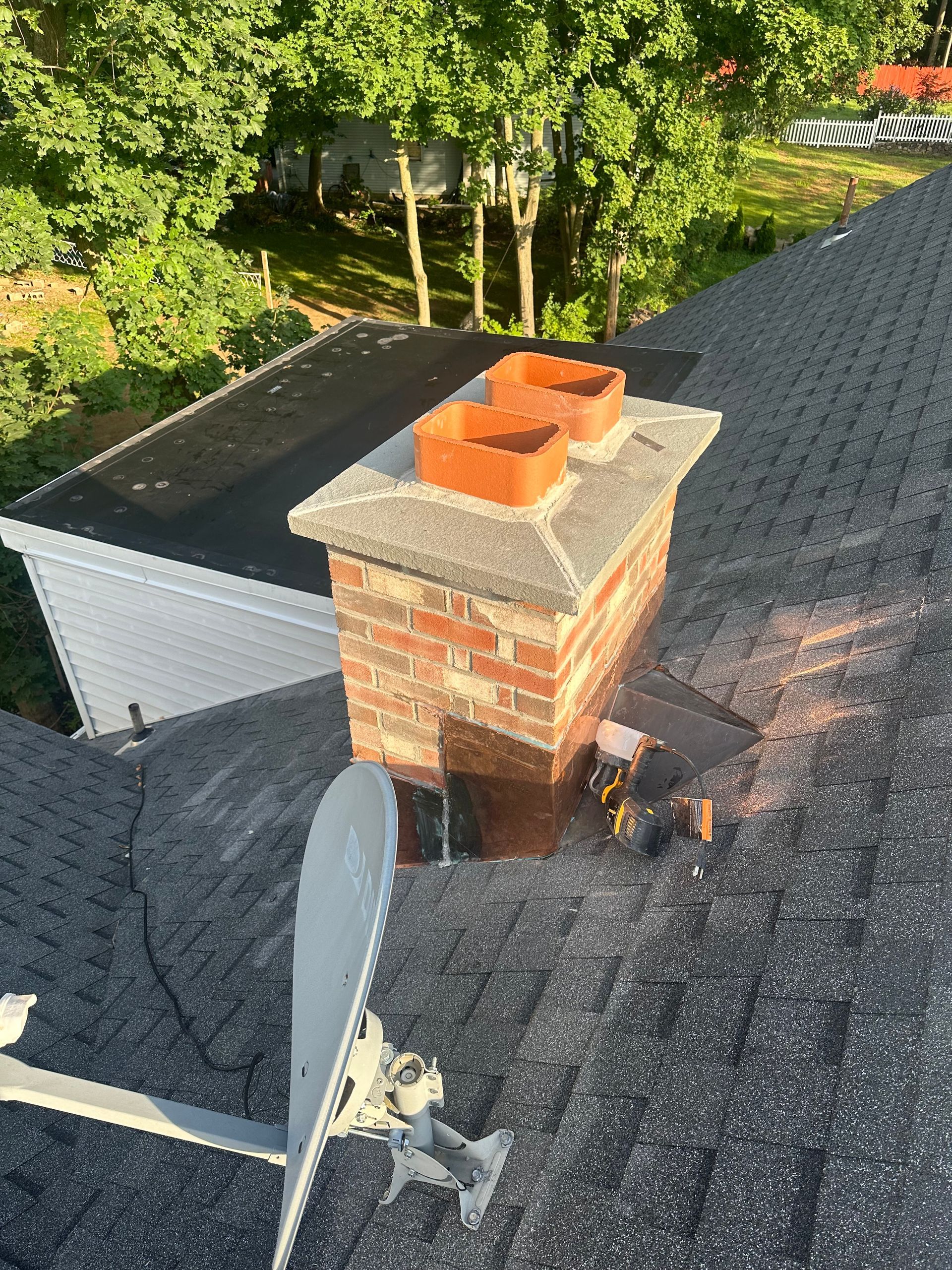 An overhead view of a brick chimney on a shingled roof, featuring two clay flue tiles and a satellite dish in the foreground.