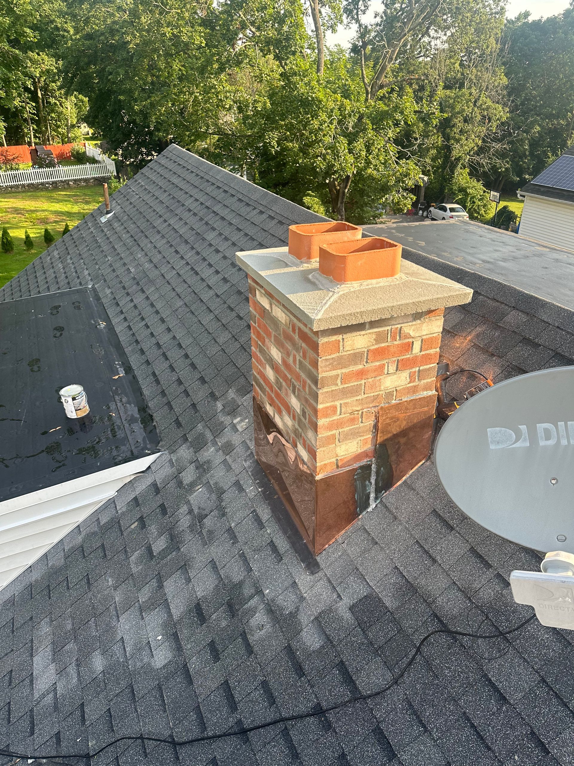 A brick chimney with two terracotta flue liners extending above a shingled roof, adjacent to a satellite dish.