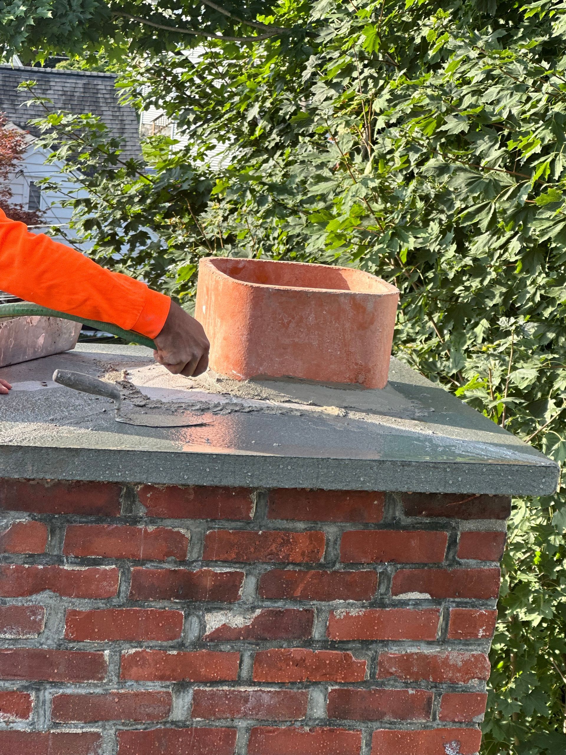 A person in an orange long-sleeved shirt uses a trowel to apply mortar around a terra cotta chimney flue on a brick chimney.