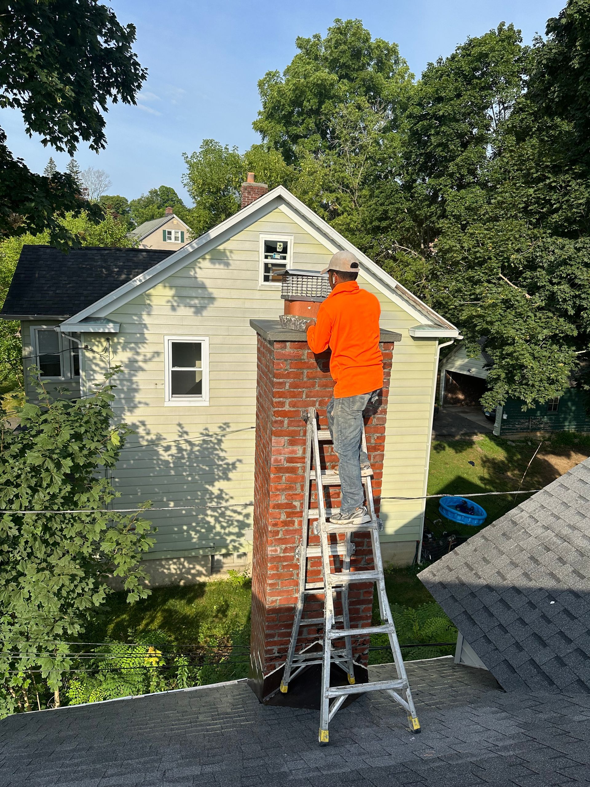 A worker in an orange shirt on a ladder repairs the top of a brick chimney against the side of a house.