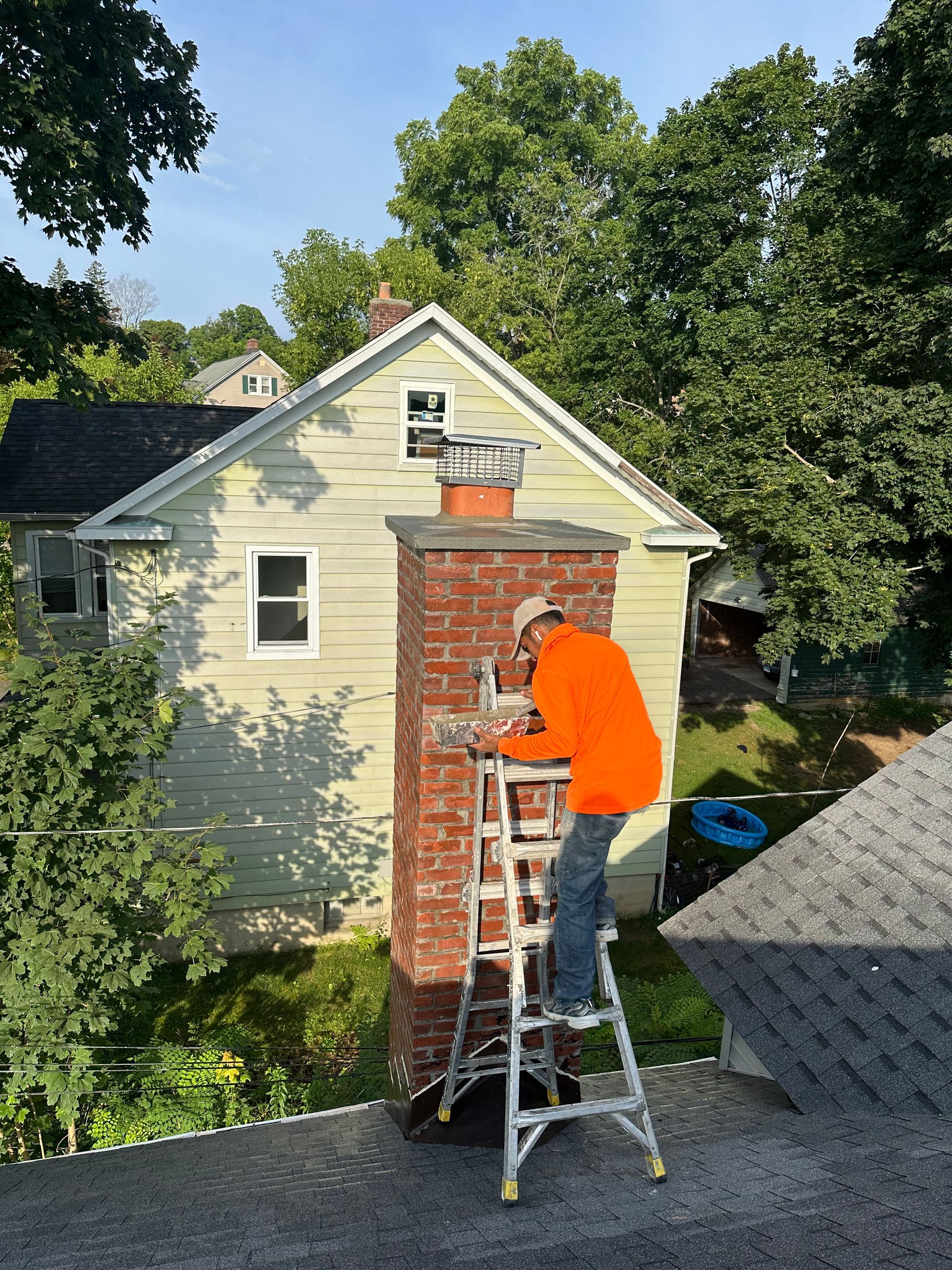 A worker in an orange sweatshirt stands on a ladder, repairing a brick chimney on a house roof.