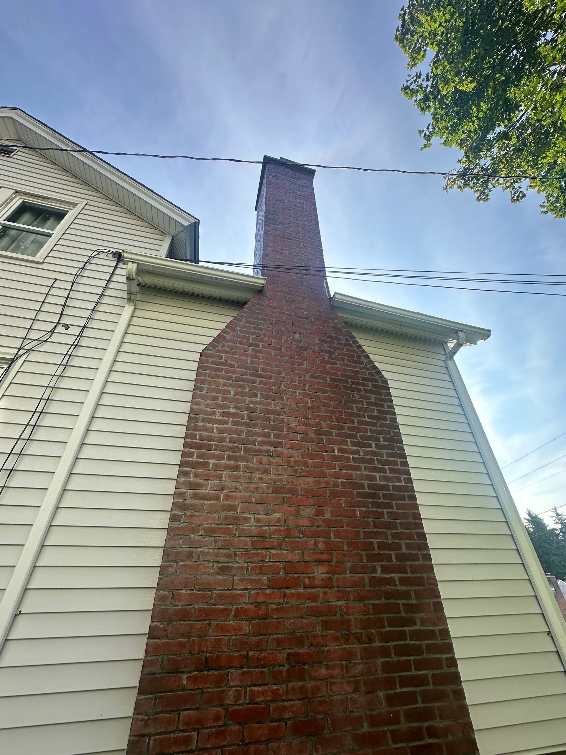 A tall, red brick chimney stands against the white-sided exterior wall of a two-story house under a blue sky.