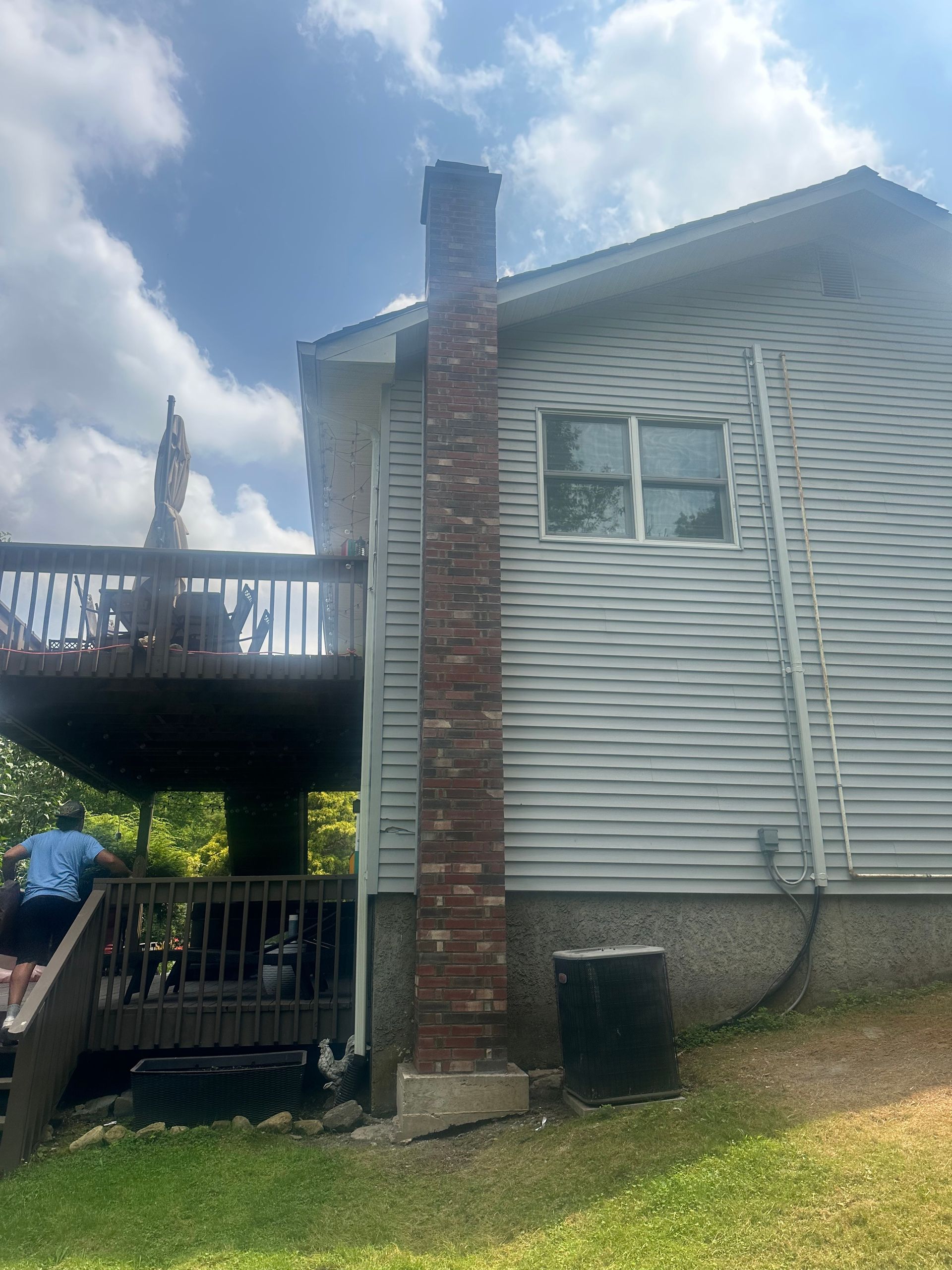 A brick chimney stands against the side of a house with grey siding, adjacent to a wooden deck.