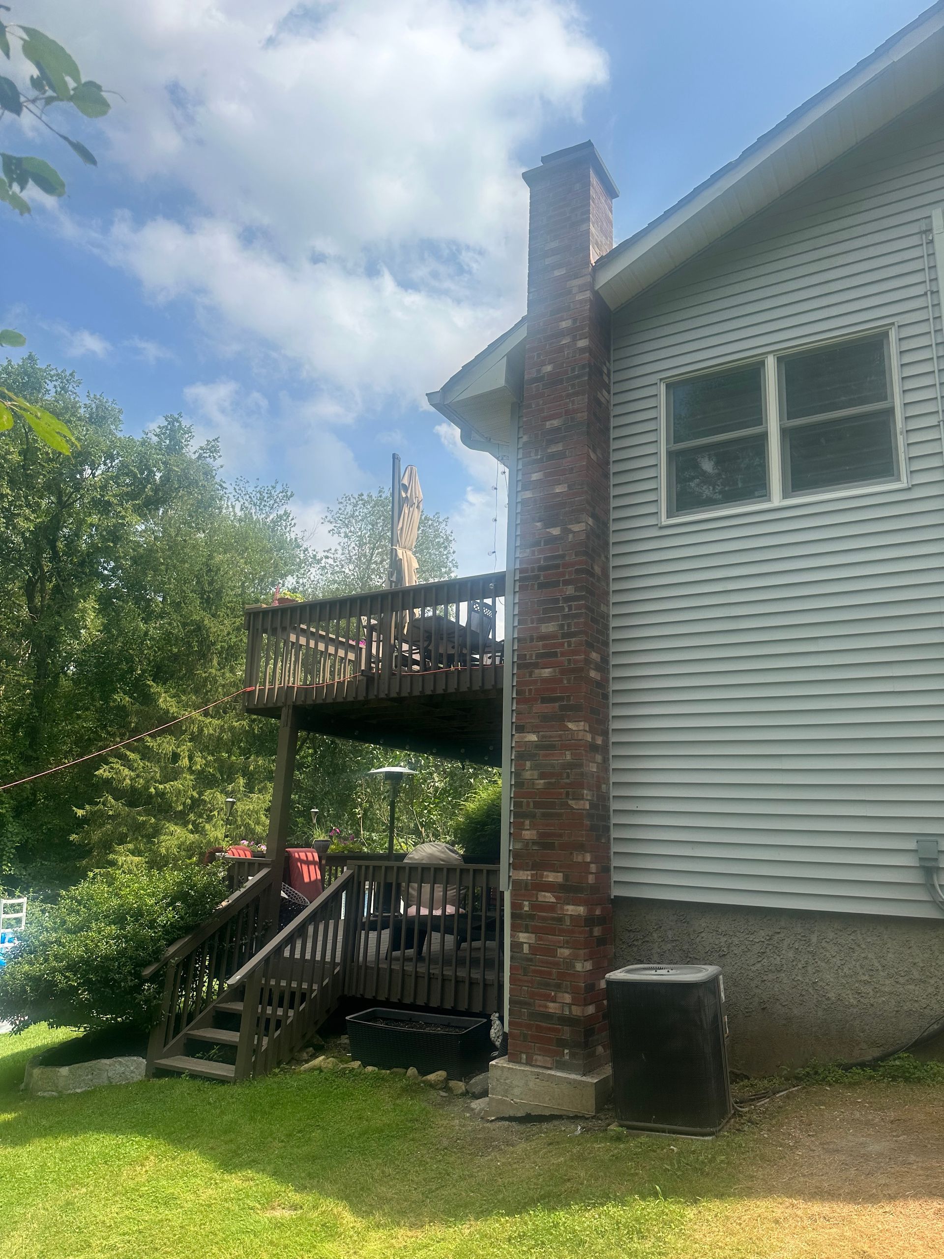 A red brick chimney stands beside a house with light gray siding, featuring a wooden deck and staircase in a backyard.