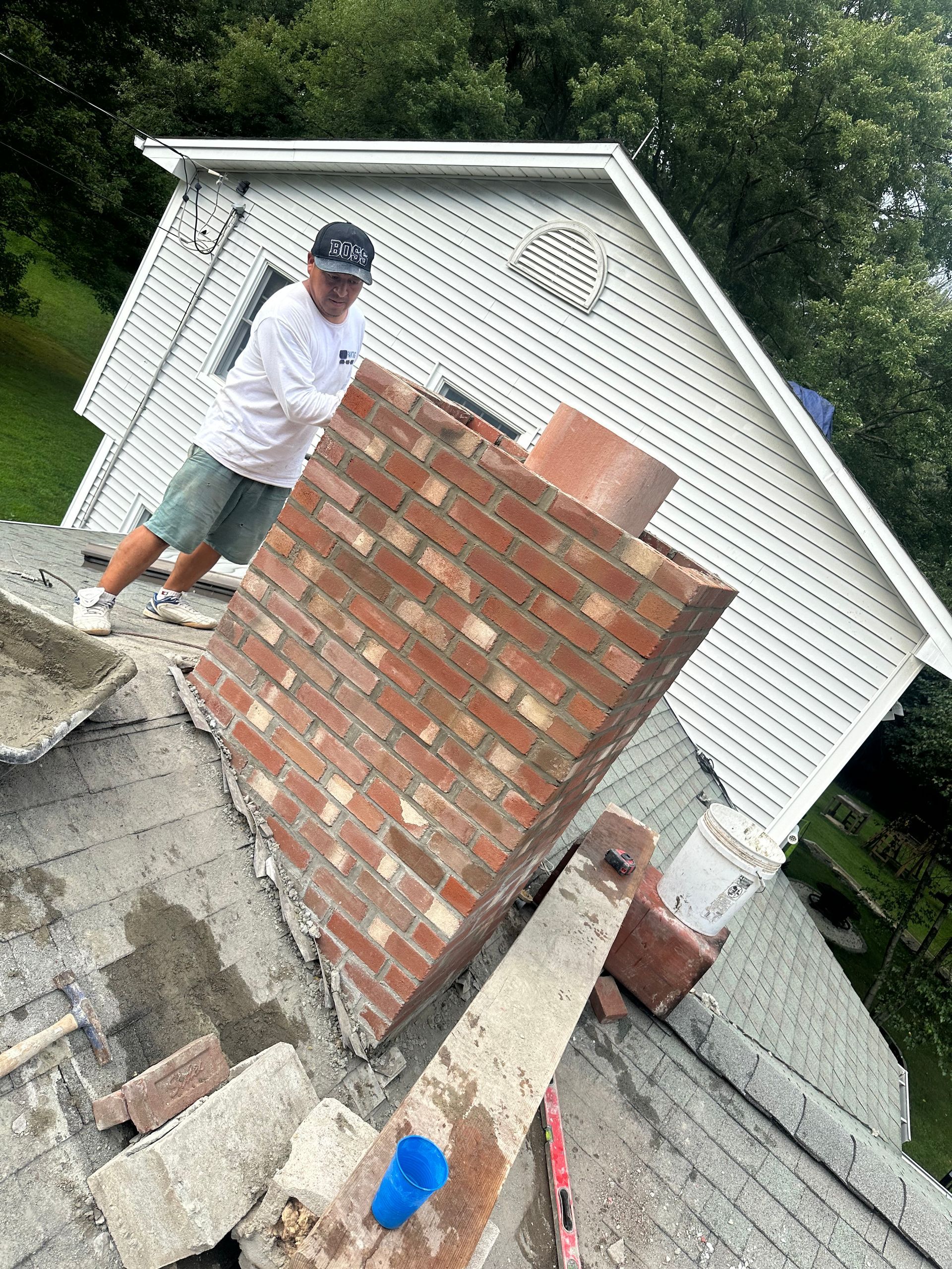 A worker building a brick chimney on a house roof next to a white lattice-sided garage.
