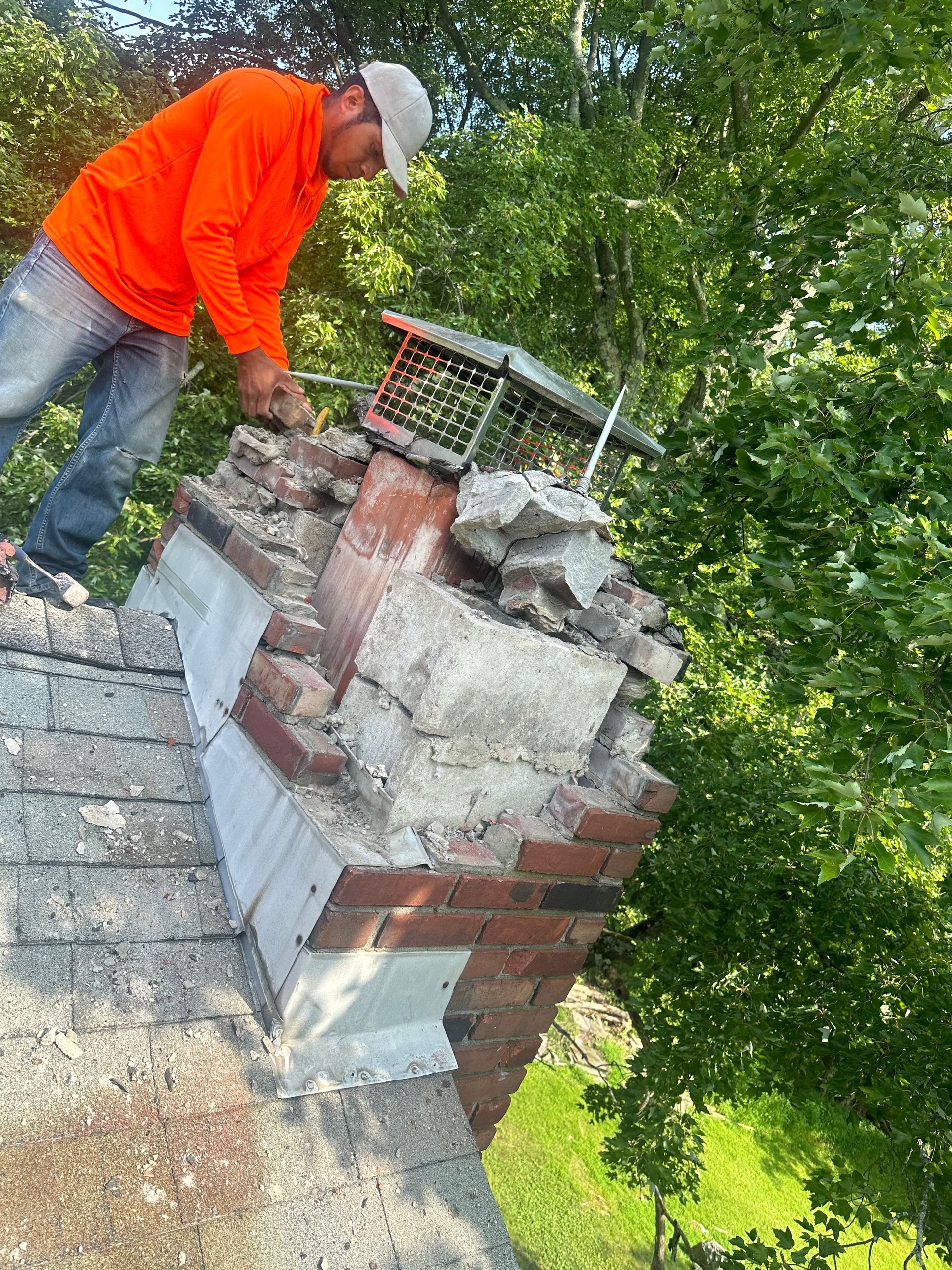A worker in an orange shirt repairs a crumbling brick chimney on a shingled roof outdoors among trees.
