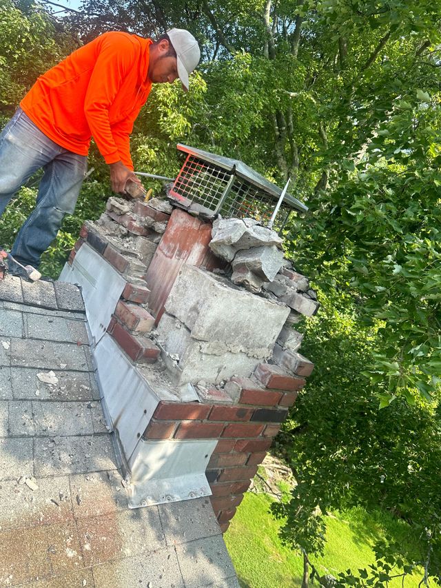 A worker in an orange long-sleeved shirt repairs a damaged brick chimney on a shingled roof surrounded by trees.