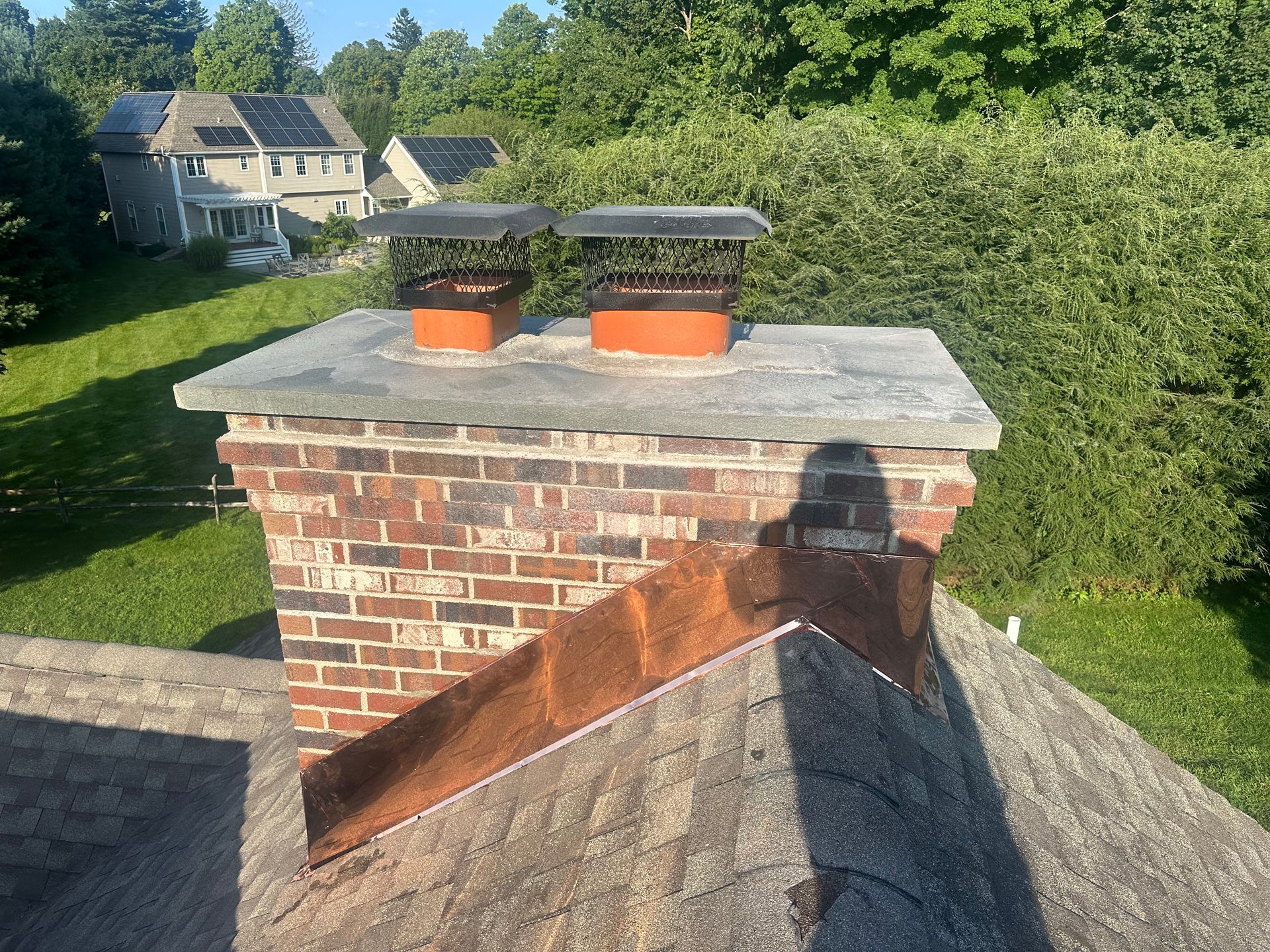 A brick chimney with two metal caps and a copper flashing base on a shingled roof under a clear blue sky.