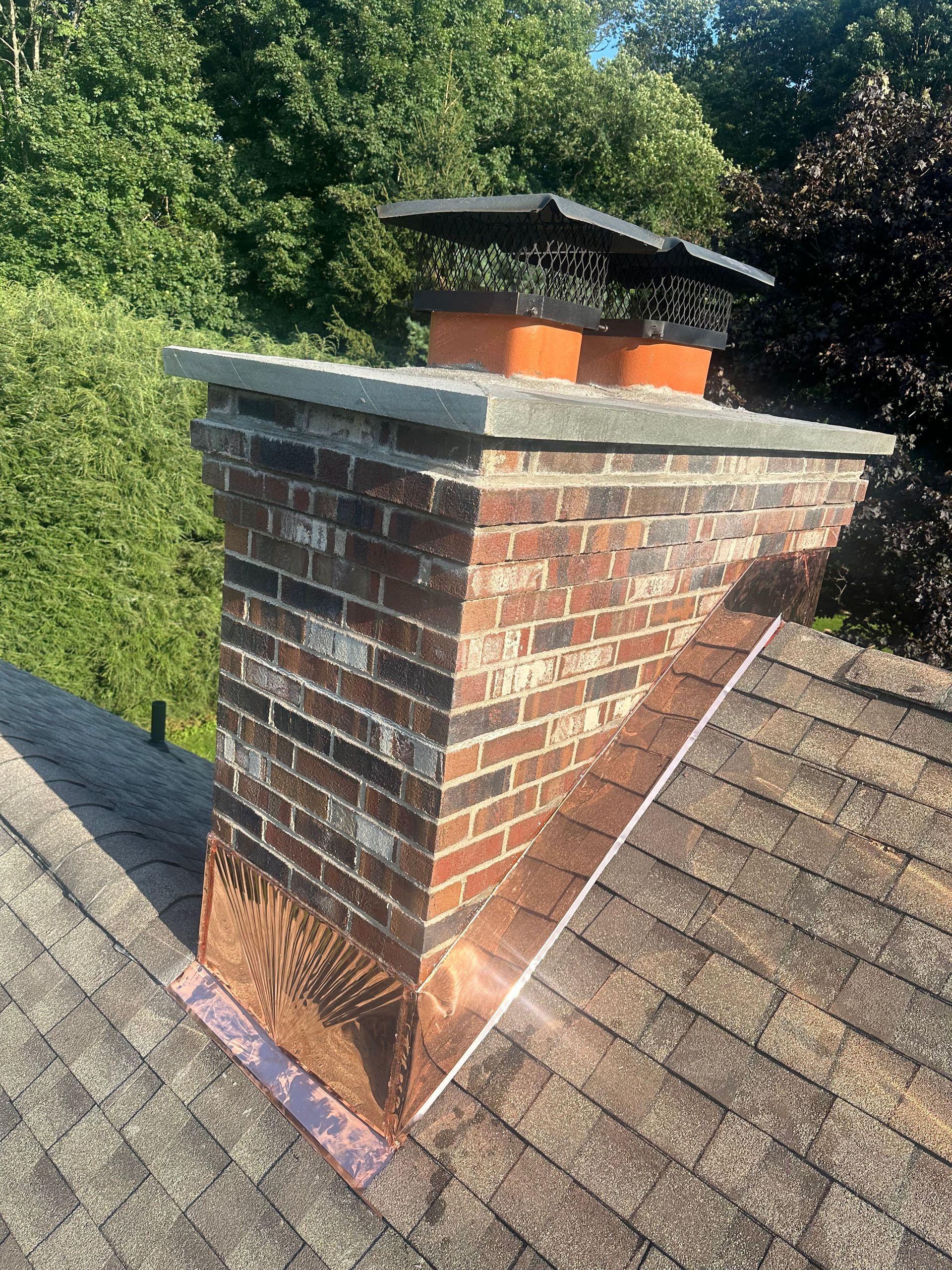 A brick chimney on a shingled roof with new copper flashing installed around its base.