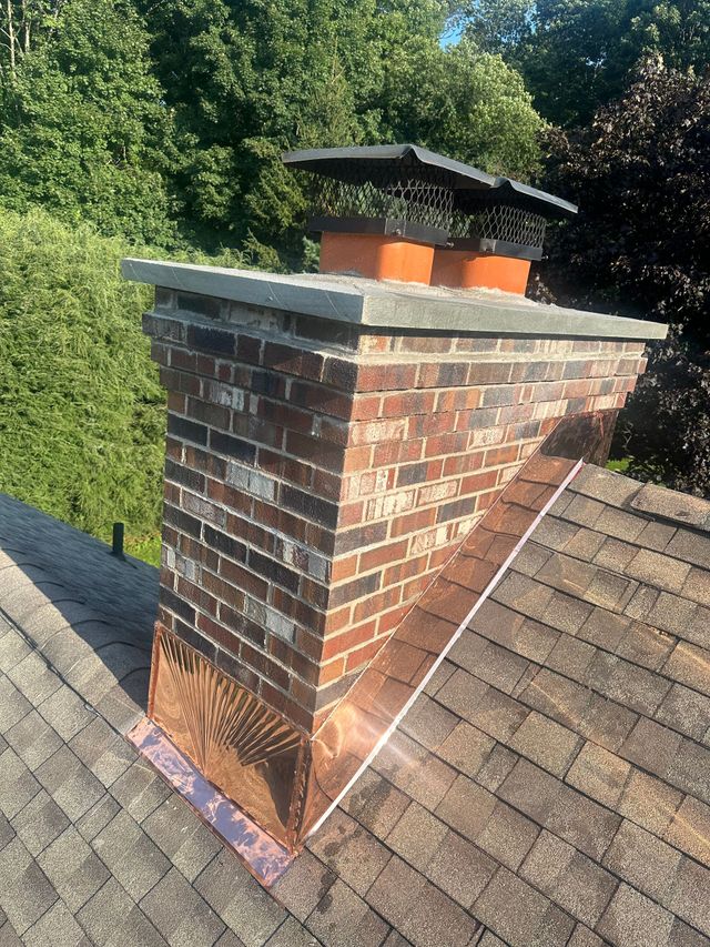 A brick chimney with a metal cap and copper flashing sits on a shingled roof under bright sunlight.