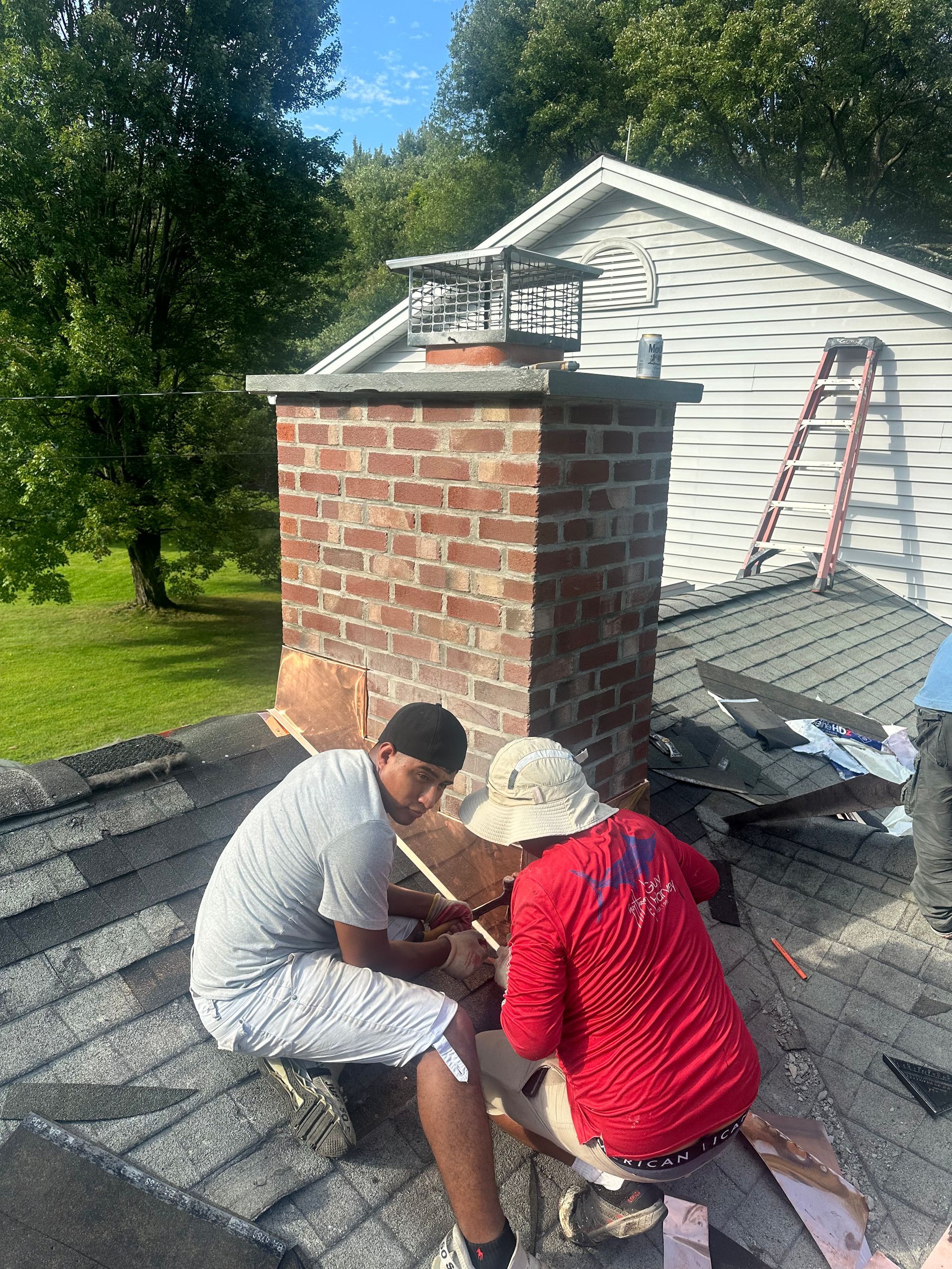 Two people in work clothes repair a brick chimney on a shingled roof under a blue sky, with a ladder visible in the back.