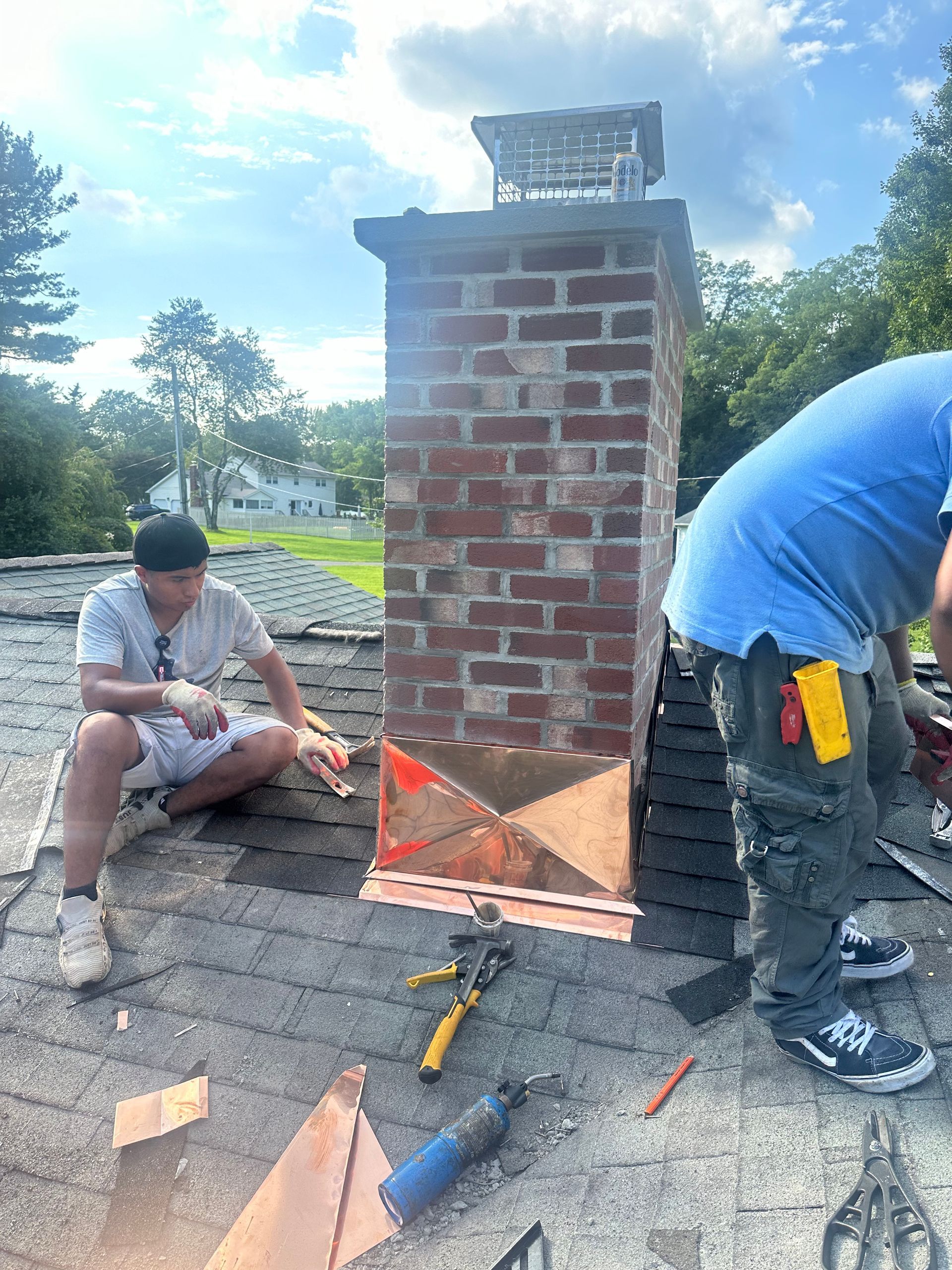 Two workers repair copper flashing at the base of a brick chimney on a shingled residential roof.