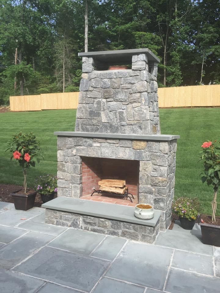 An outdoor stone fireplace sits on a gray stone patio in a grassy backyard, flanked by two potted plants.