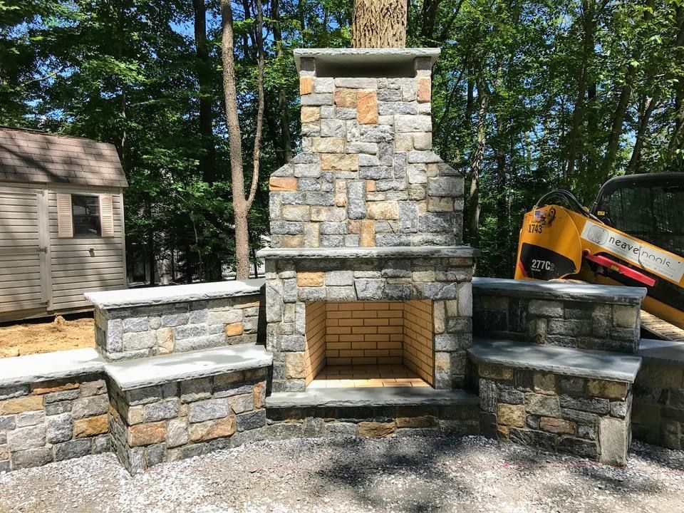 A stone outdoor fireplace with a brick interior and matching tiered side benches set in a wooded backyard.