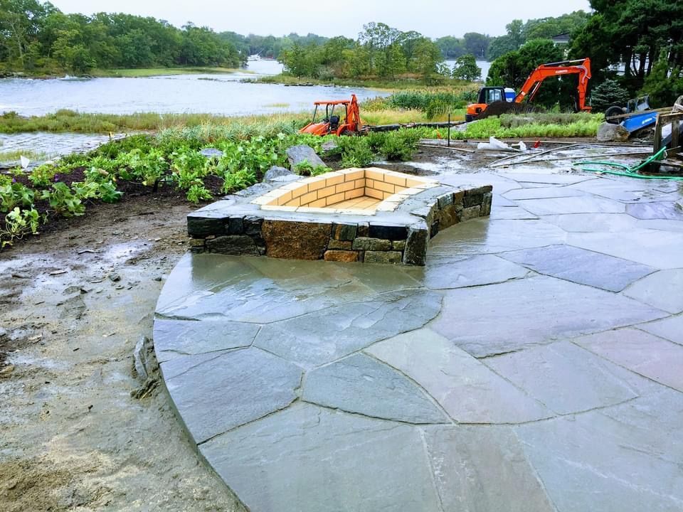 A stone fire pit sits on a large slate patio under construction, with a body of water and heavy machinery in the background.