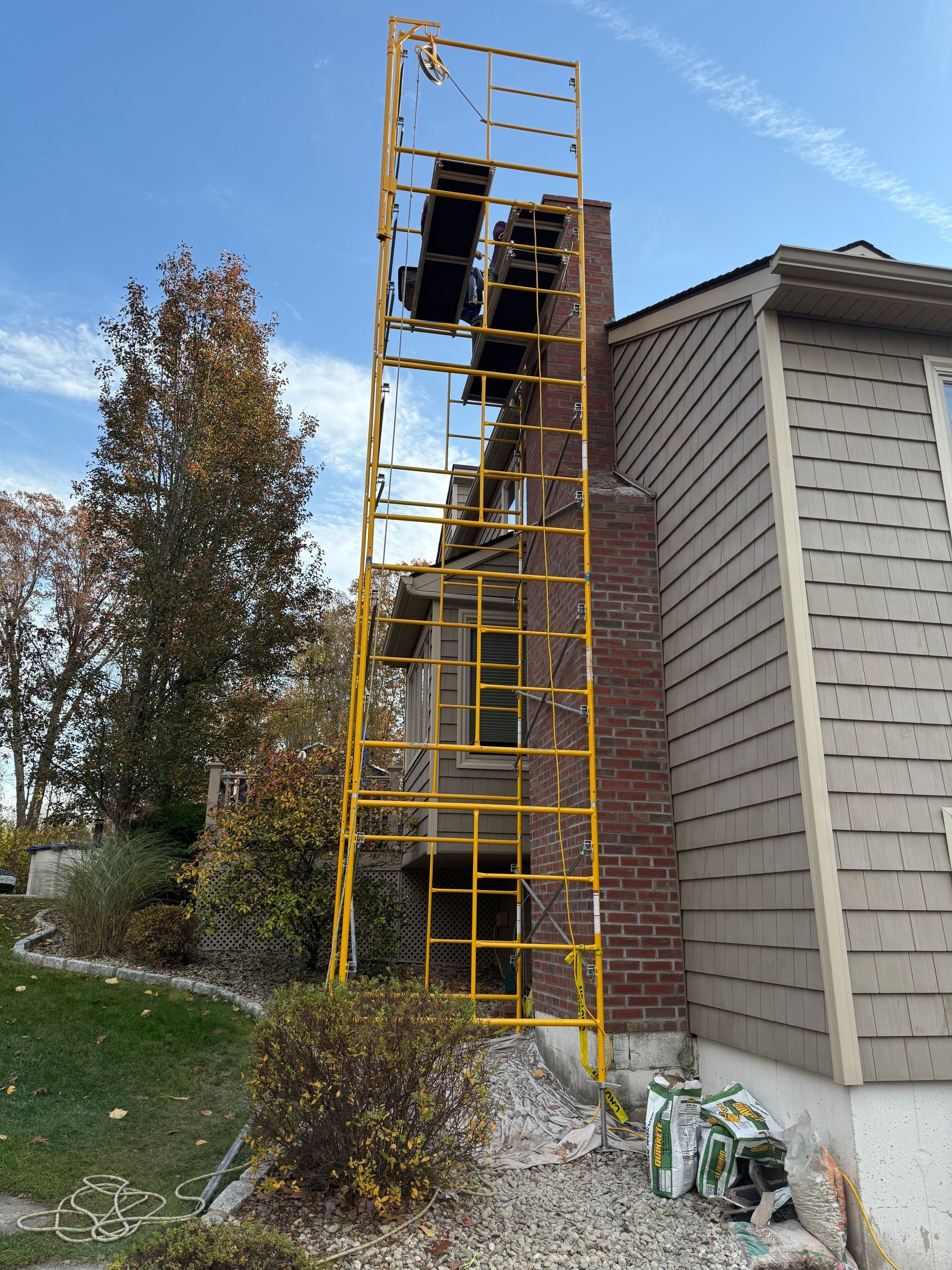 Yellow scaffolding standing against a brick chimney on the side of a suburban house with gray siding.