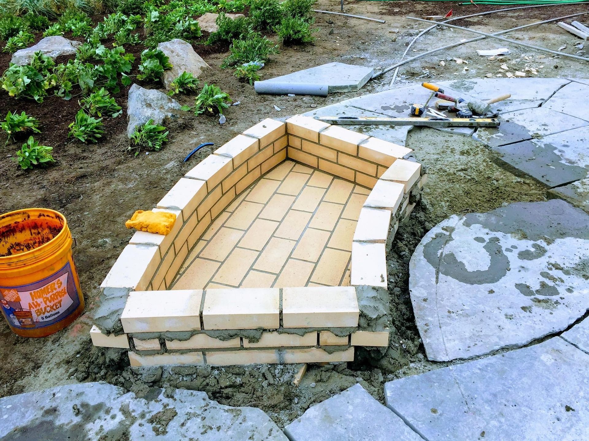 A partially built rectangular fire pit made of light-colored bricks sits on a stone patio under construction.