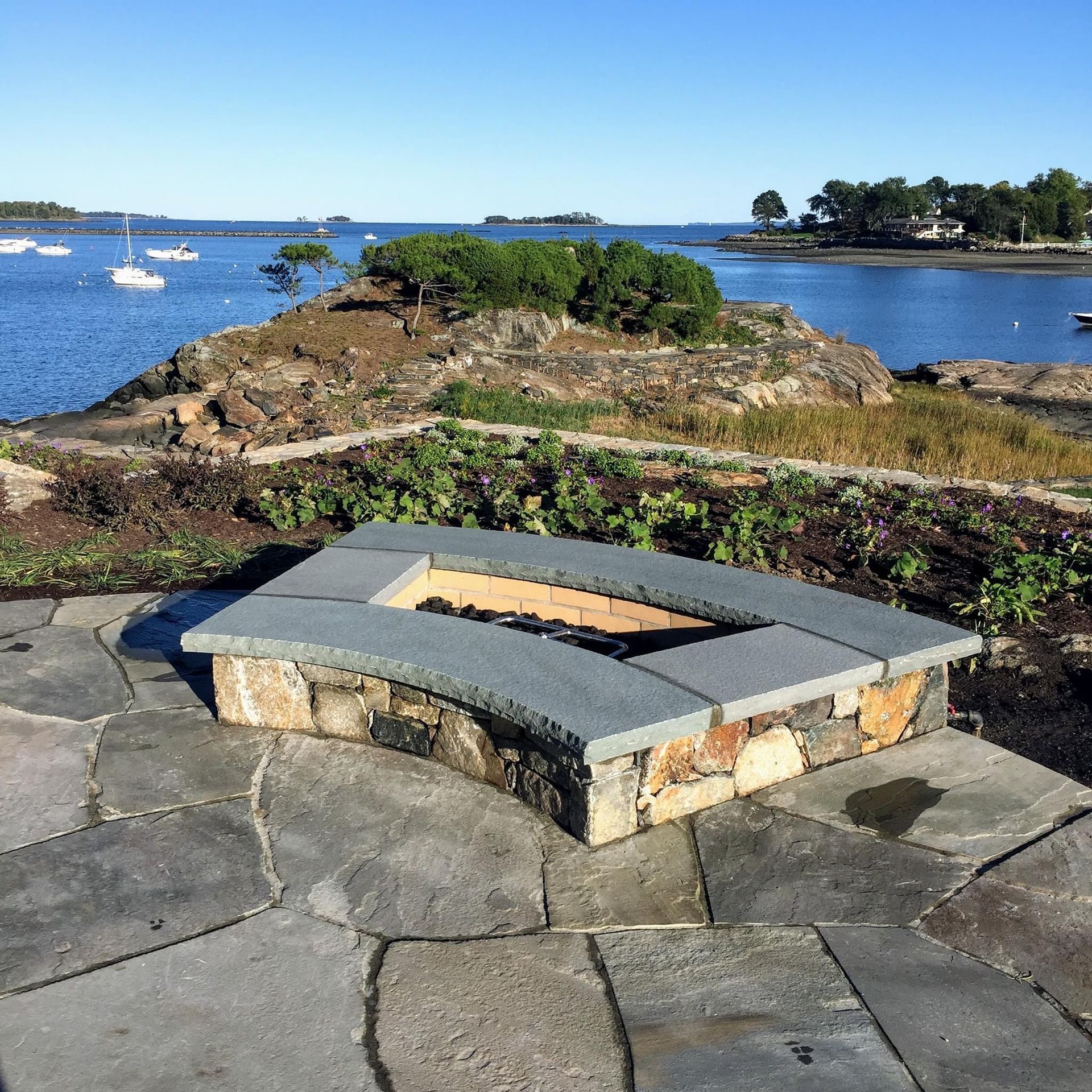 A stone fire pit on a flagstone patio overlooking a bay with boats, a rocky island, and trees under a clear blue sky.