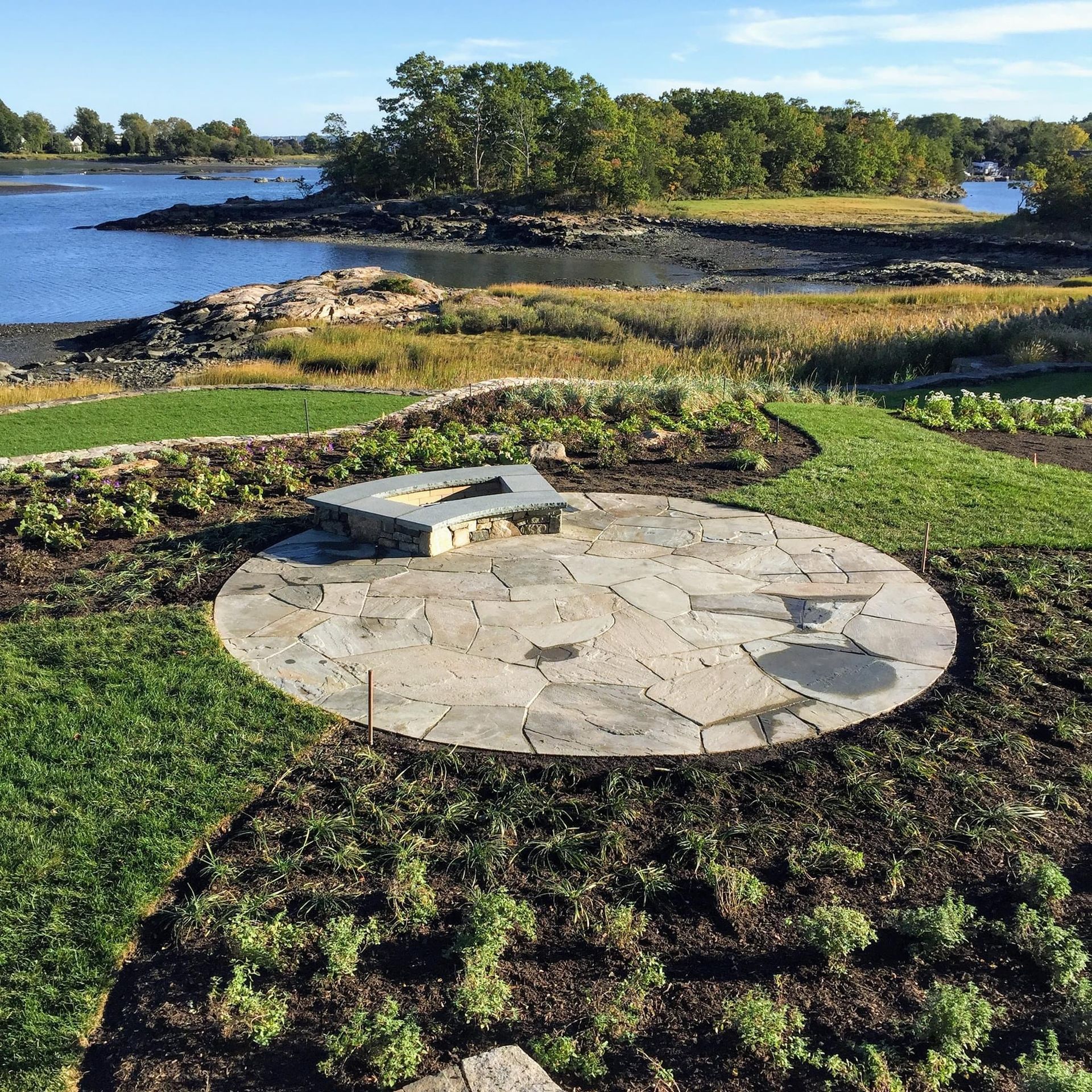 A stone fire pit sits on a circular flagstone patio overlooking a tidal inlet surrounded by green lawn and vegetation.