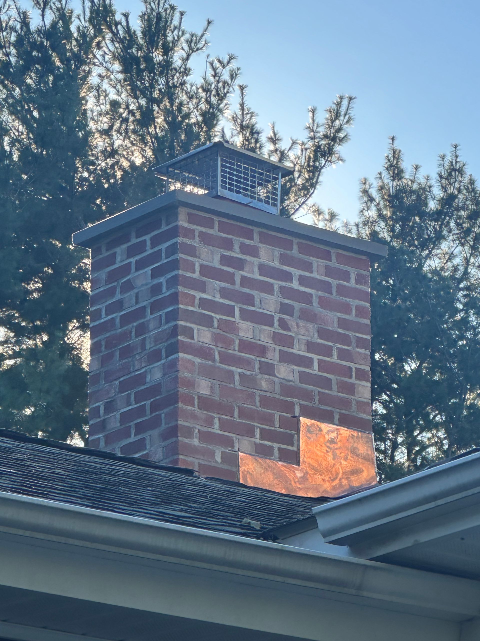 A red brick chimney with a metal cap and copper flashing stands on a roof against a backdrop of trees.