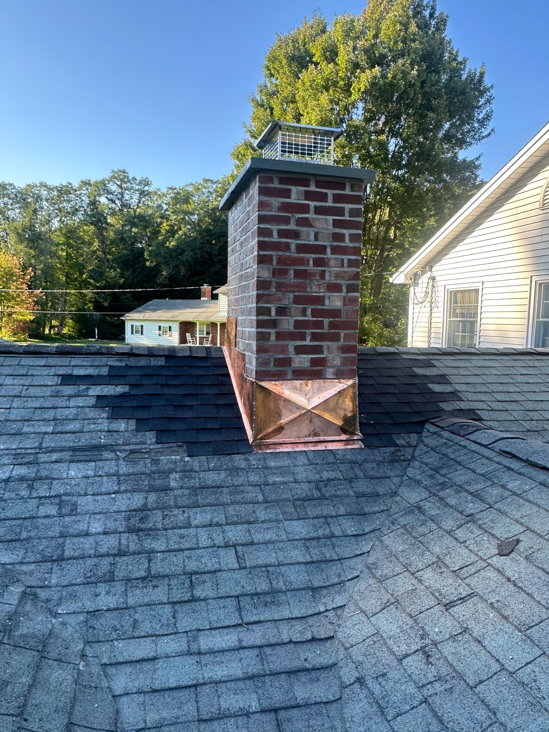 A brick chimney on a shingled roof with a copper cricket installed at the base, set against a backdrop of trees and a house.
