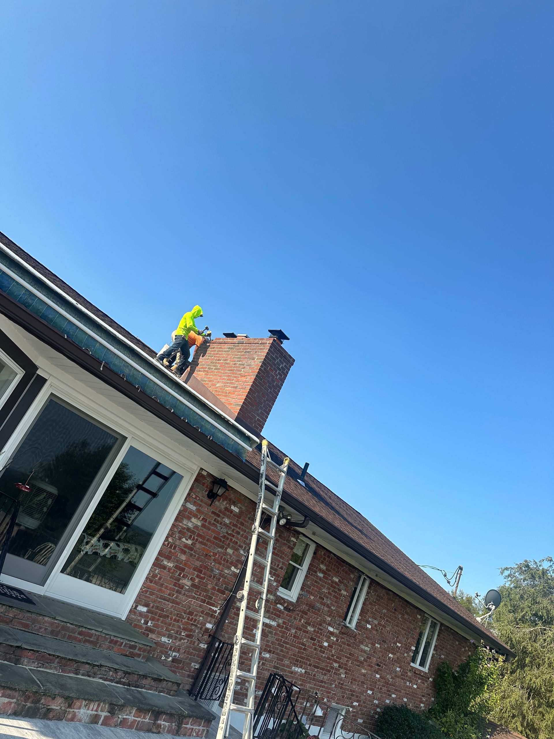 A worker in a bright yellow safety jacket stands on a residential brick roof next to a chimney, accessed by a tall ladder.