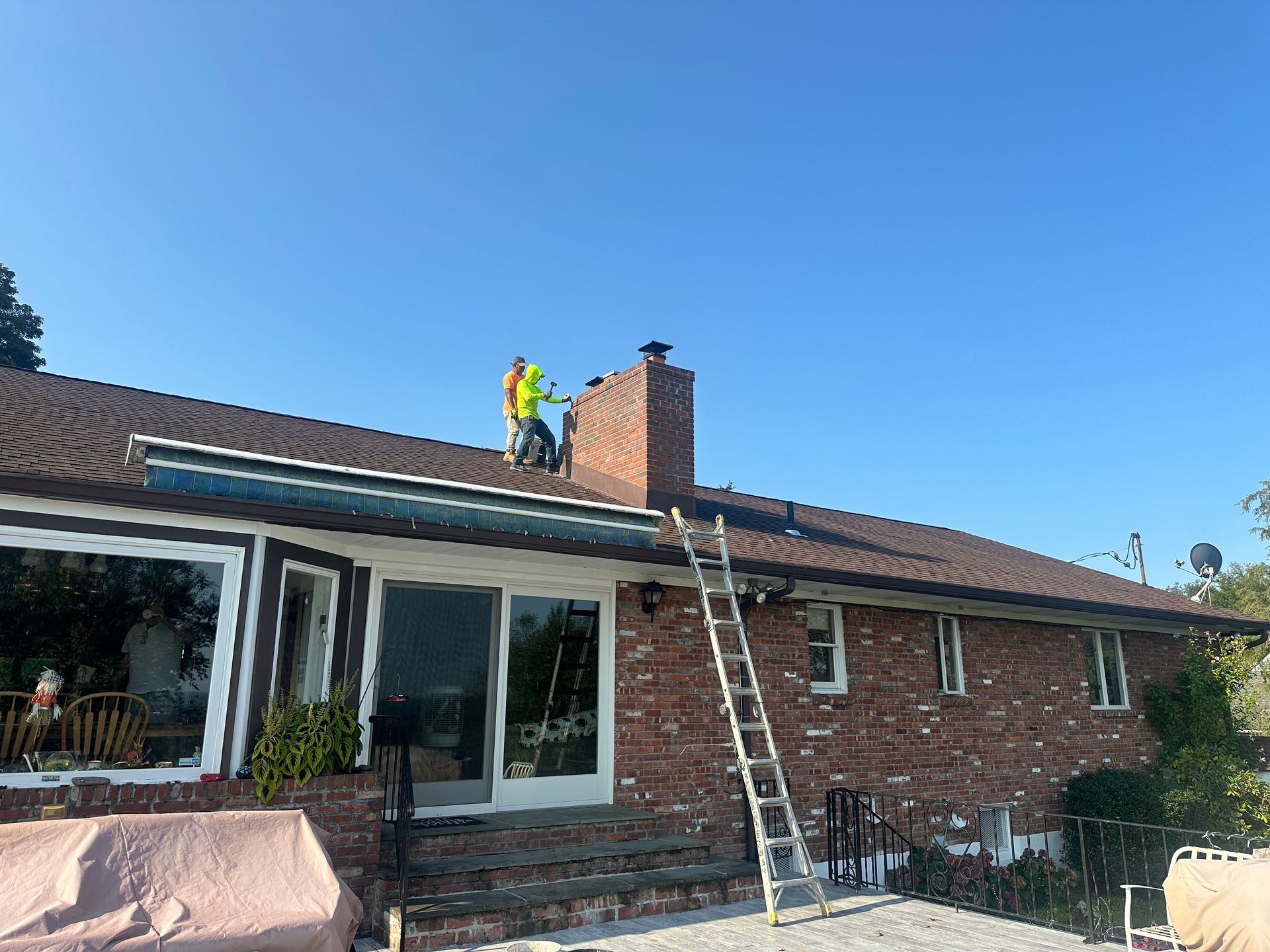 A worker in a bright yellow safety vest stands on a brown roof near a brick chimney, with a ladder leaning against it.