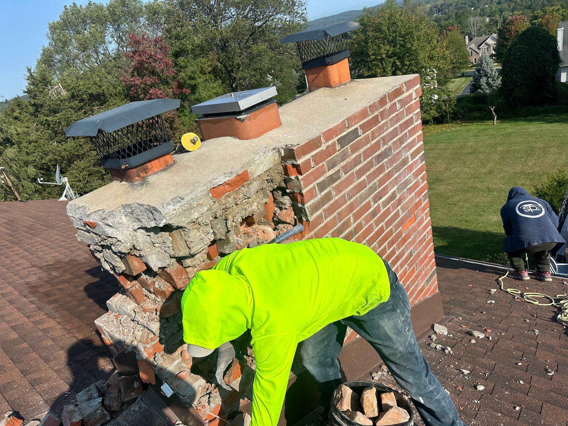 A worker in a bright yellow hoodie repairs a damaged brick chimney on a sloped roof while another person works nearby.