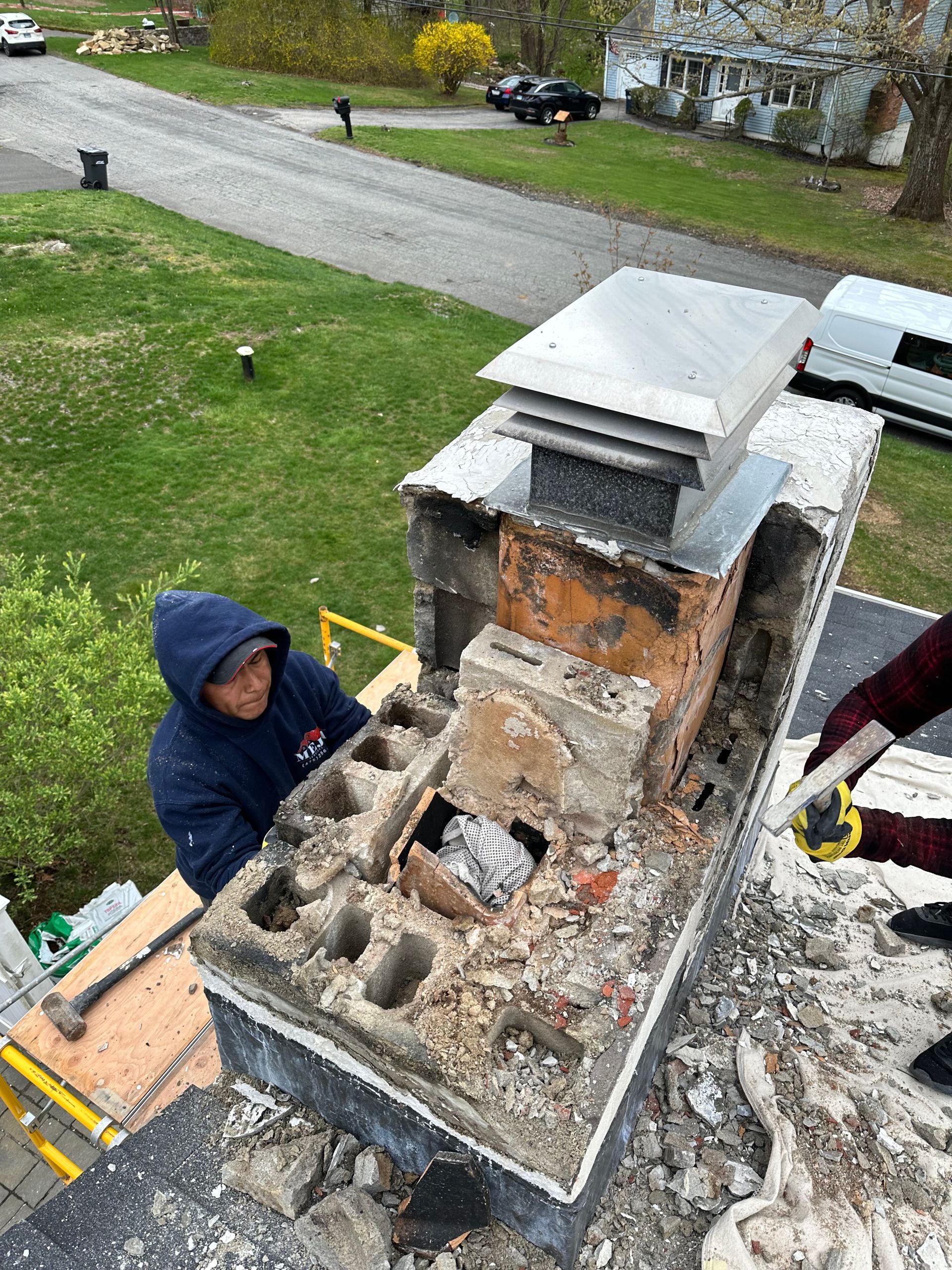 A worker in a blue hoodie repairs a damaged brick chimney on a rooftop, surrounded by debris and construction tools.