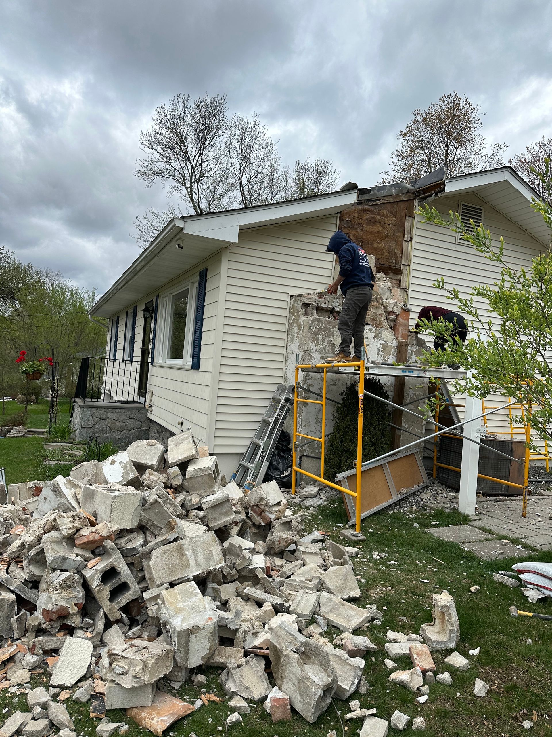A person on scaffolding works on a home's exterior, removing masonry from a chimney with a large pile of rubble below.