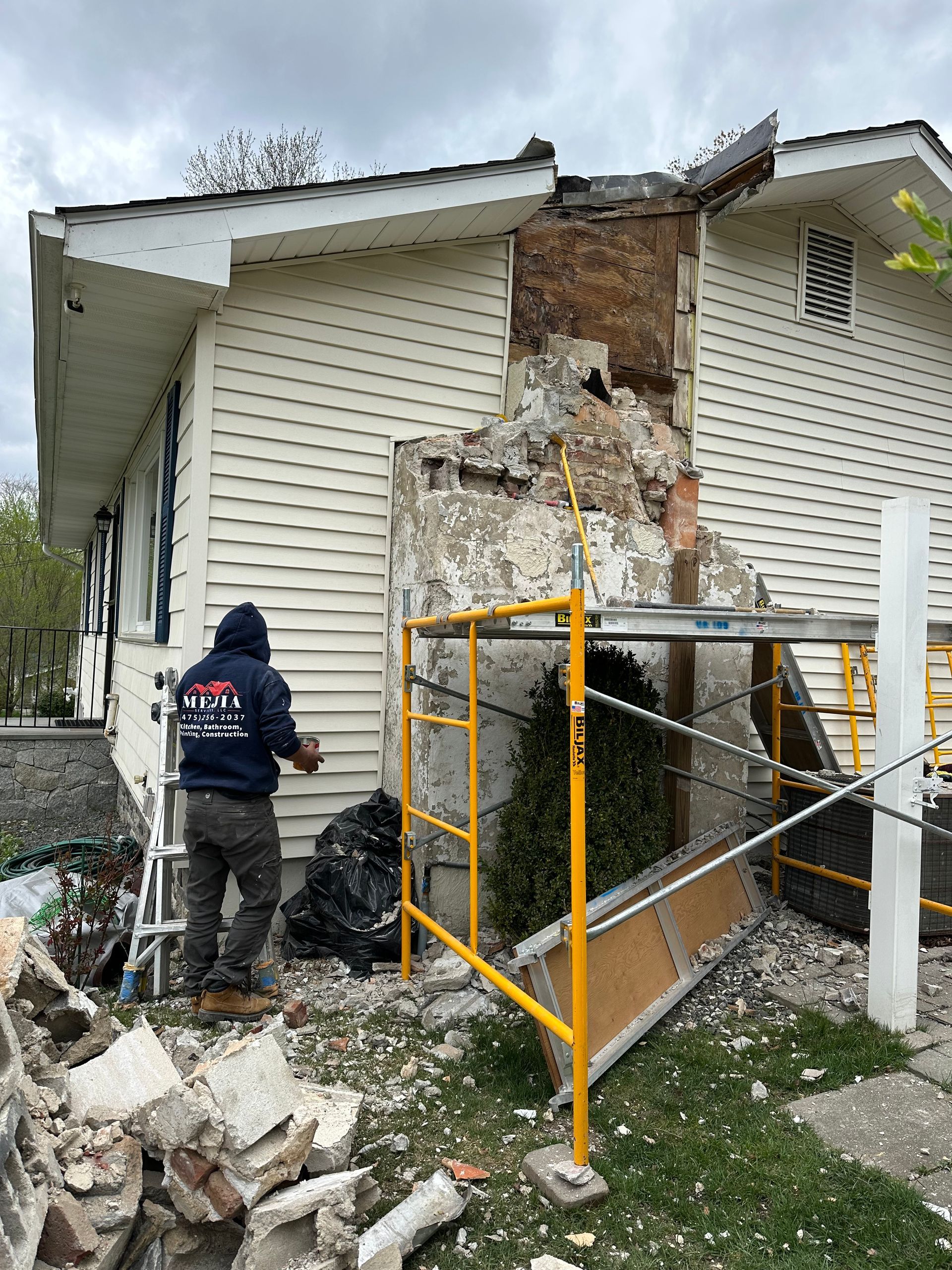 A person in a blue hoodie works on dismantling a brick chimney on the side of a white house, using yellow scaffolding.