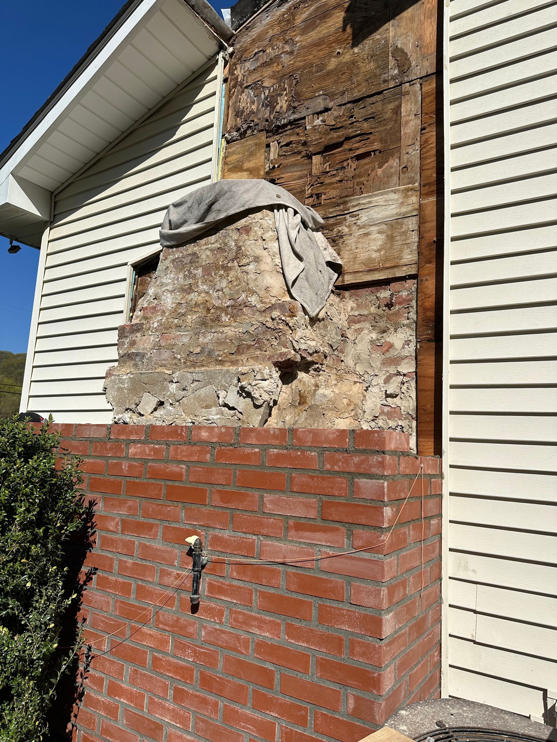 A partially demolished brick fireplace exterior with exposed wall structure and siding on the side of a house.