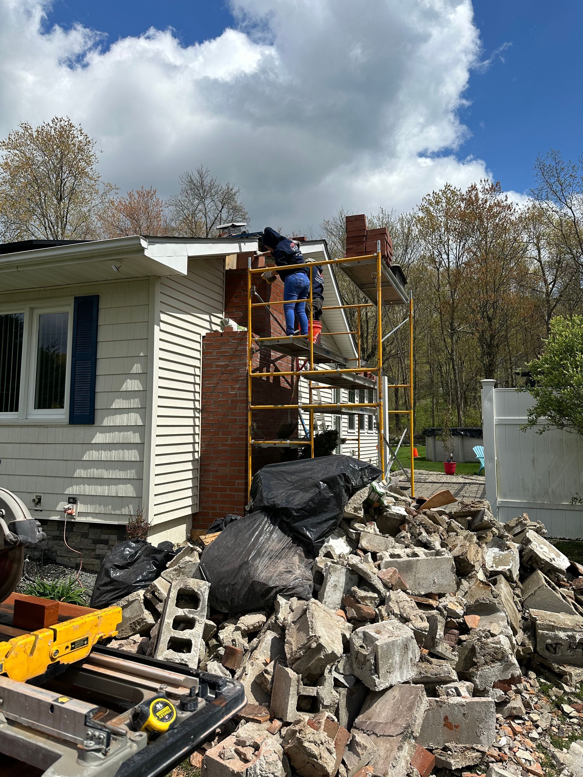 A person on scaffolding works to repair a brick chimney on a house, surrounded by a pile of rubble and debris.