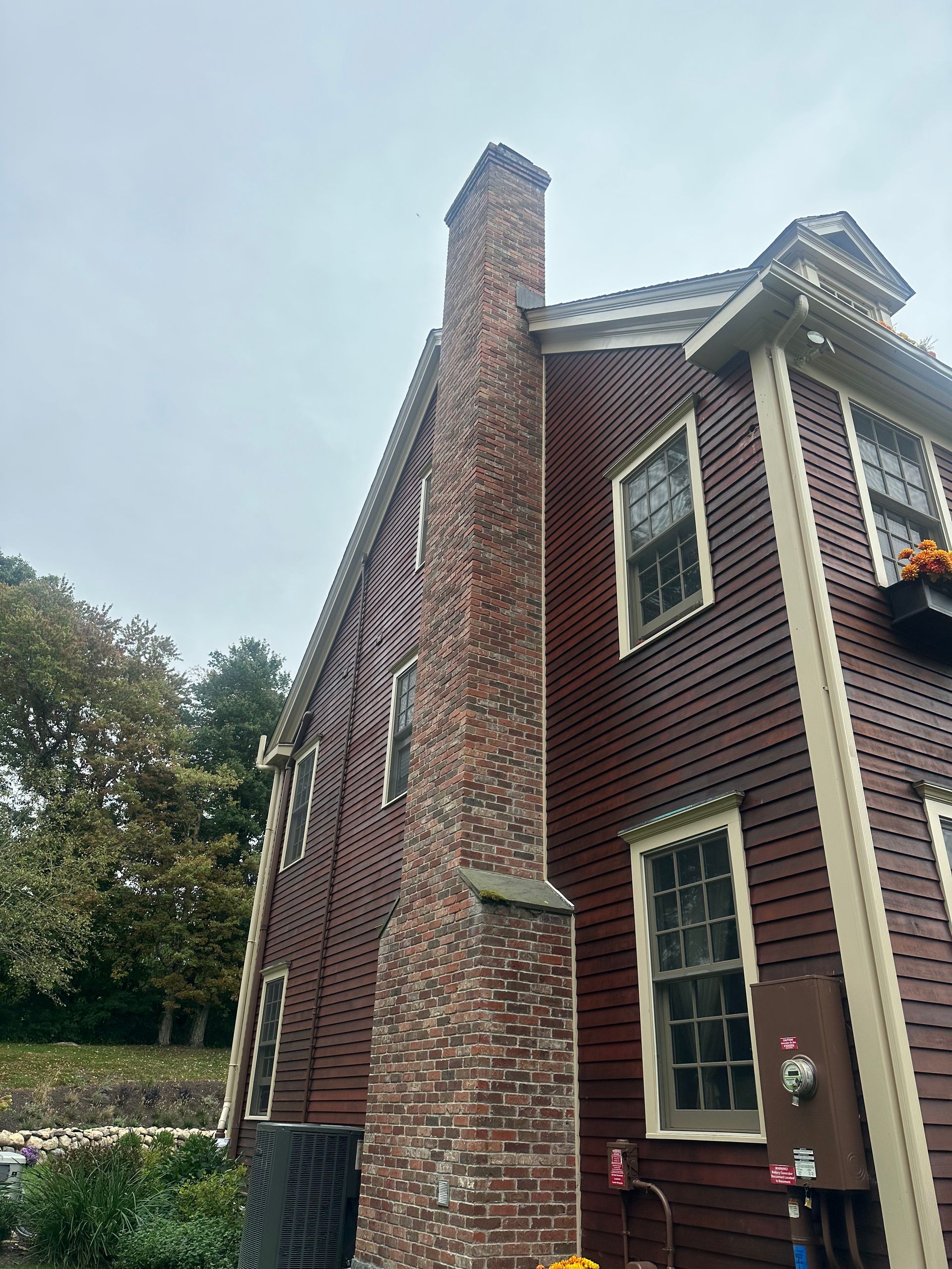 A side view of a red-sided house with a large brick chimney, white trim, and multi-pane windows under a cloudy sky.