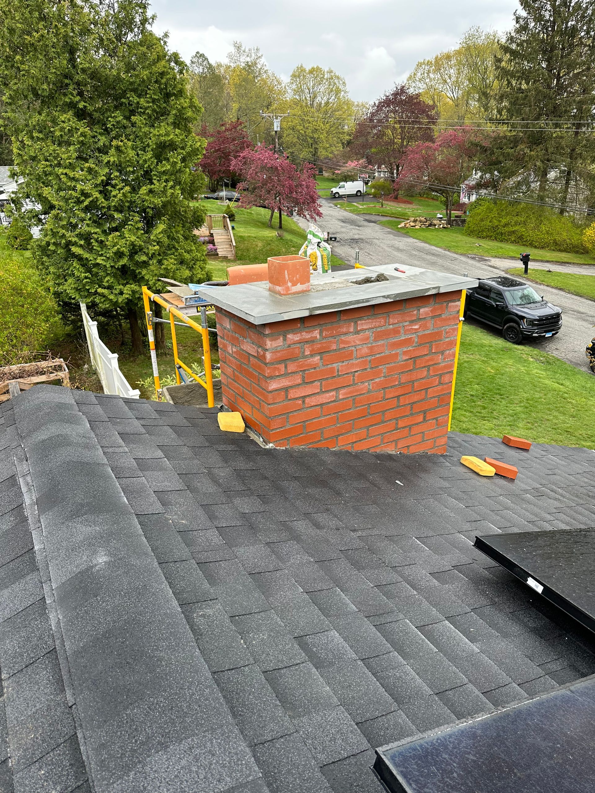 A red brick chimney with a gray stone cap on a black shingled roof, with yellow scaffolding visible in the background.