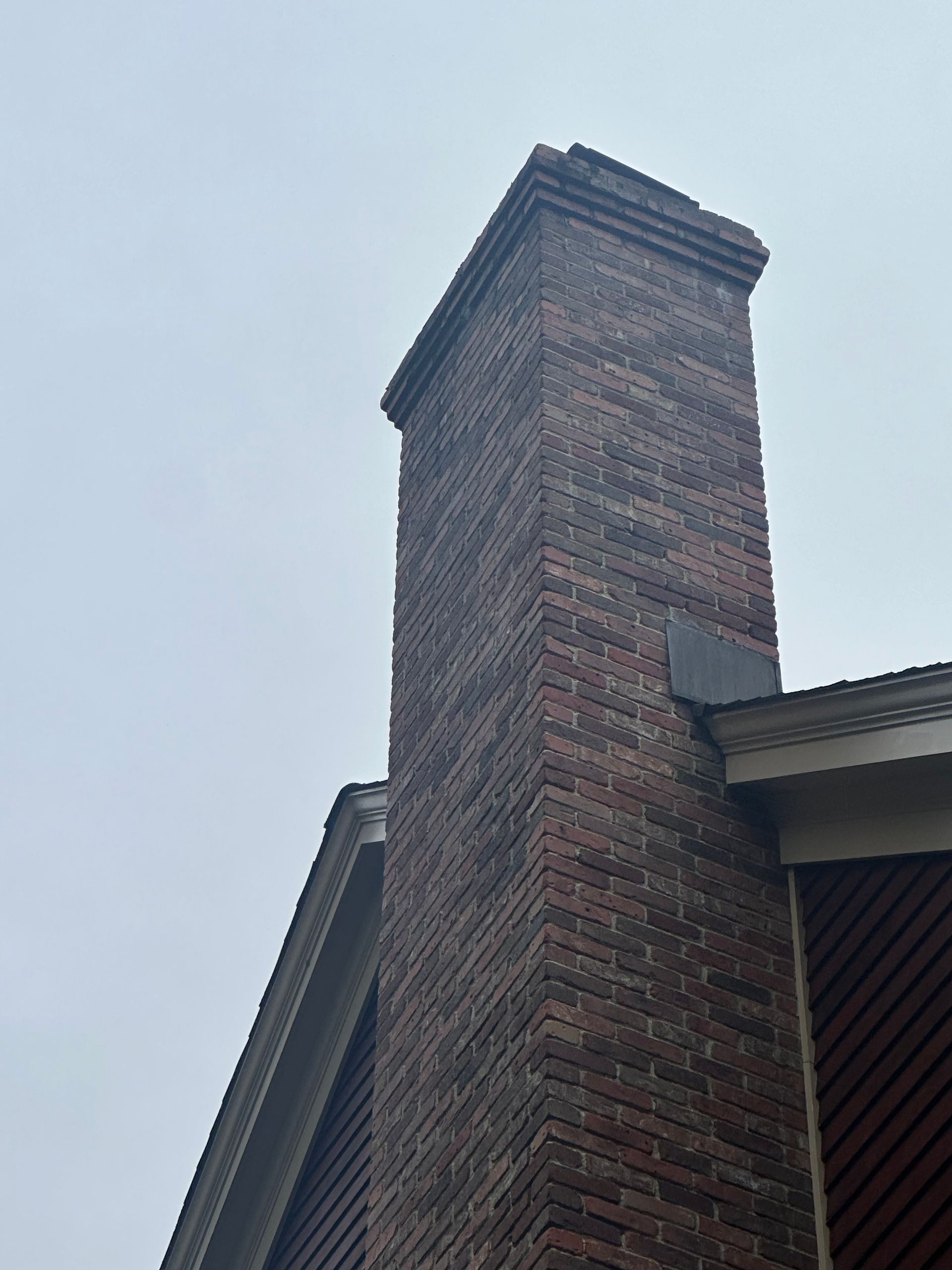 A brick chimney rises against a cloudy sky, positioned next to the eaves and siding of a house.