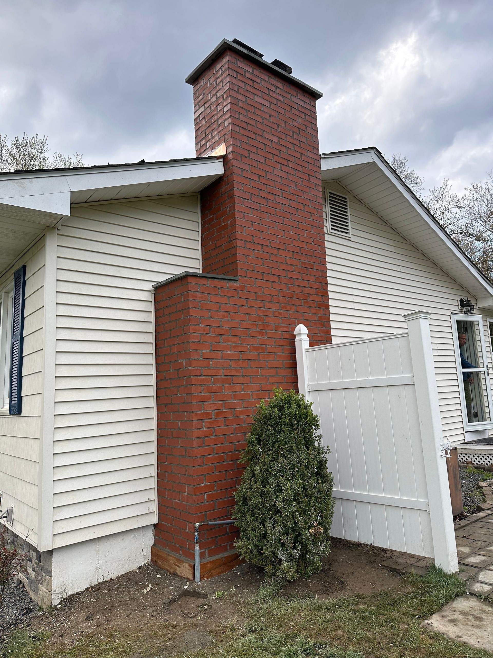 A red brick chimney rises against the exterior of a white, vinyl-sided house next to a white privacy fence and a shrub.