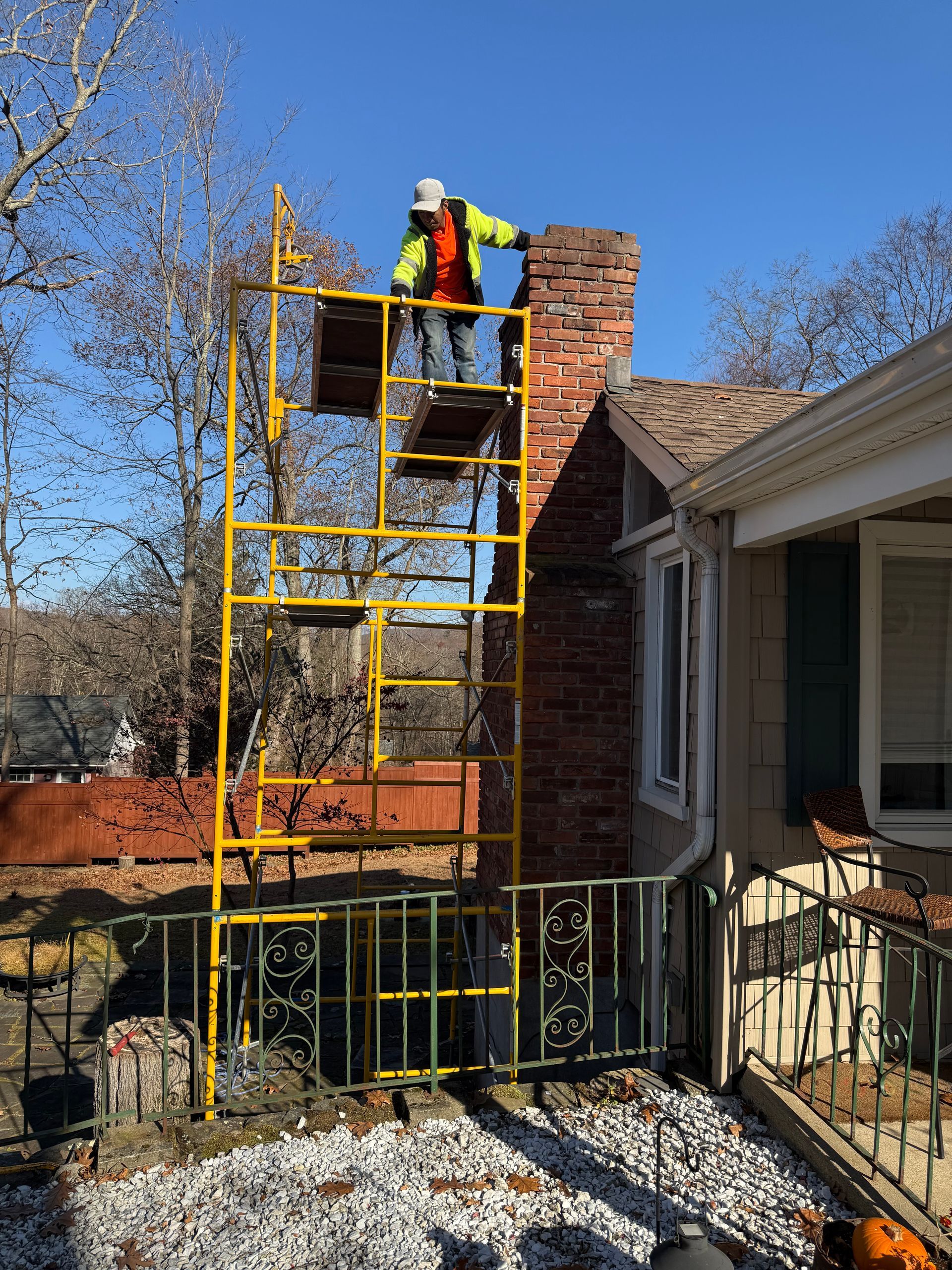 A worker in a high-visibility jacket stands on yellow scaffolding to repair a brick chimney on a house exterior.