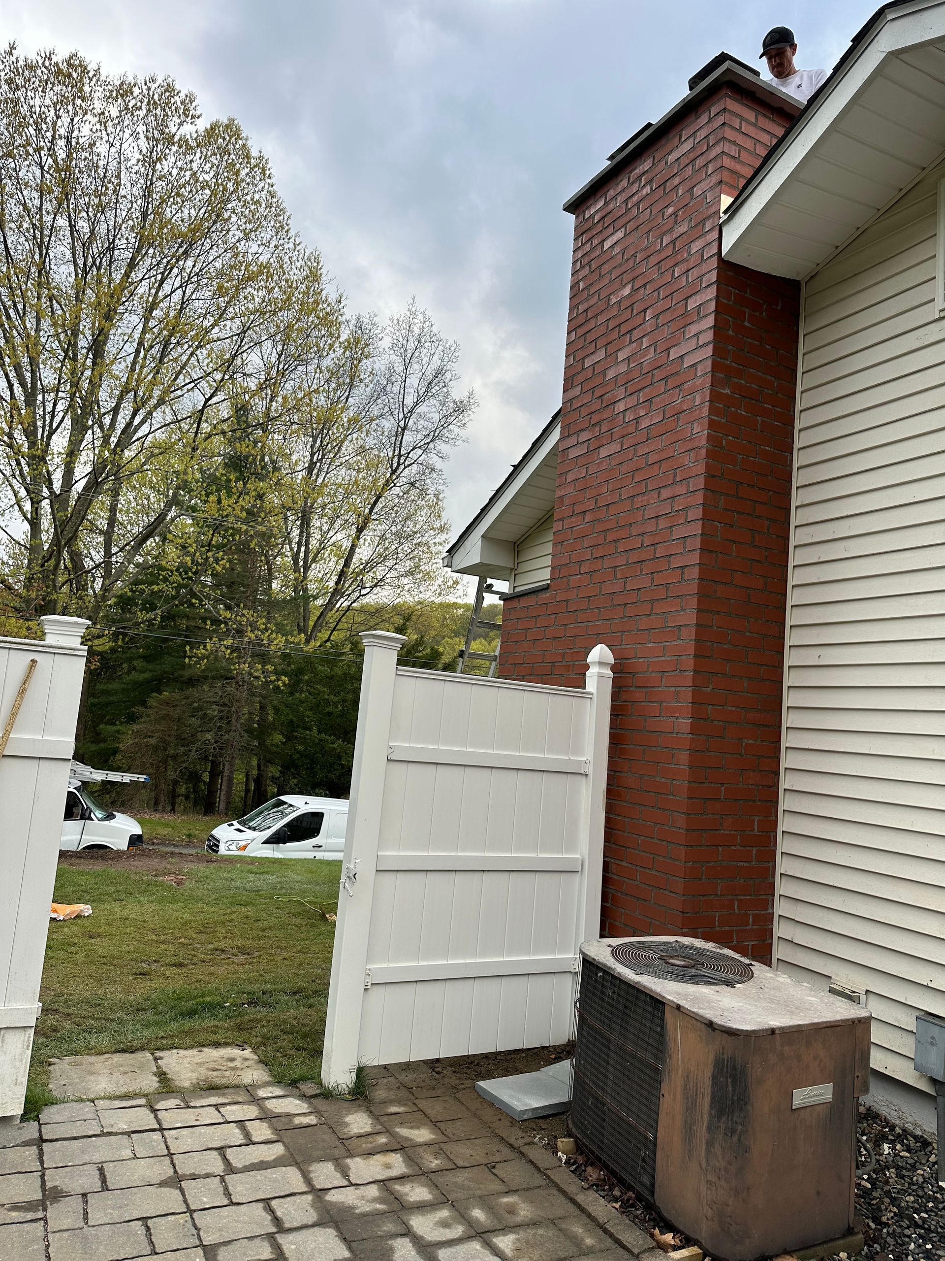 A person on a roof inspecting a red brick chimney next to a white house, a privacy fence, and an outdoor HVAC unit.