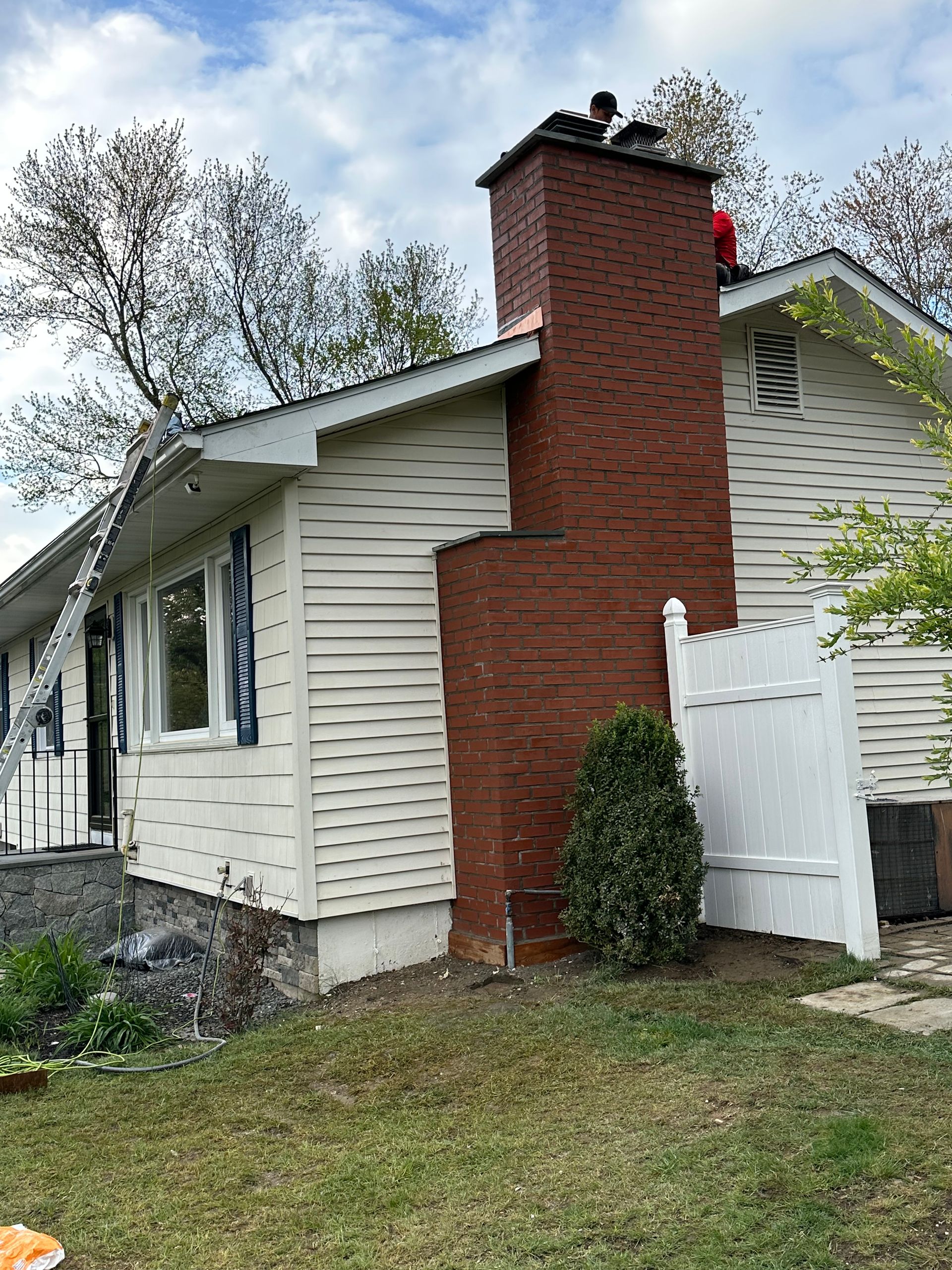 A brick chimney on a house with light-colored siding, with a ladder leaning against the side and a person on the roof.