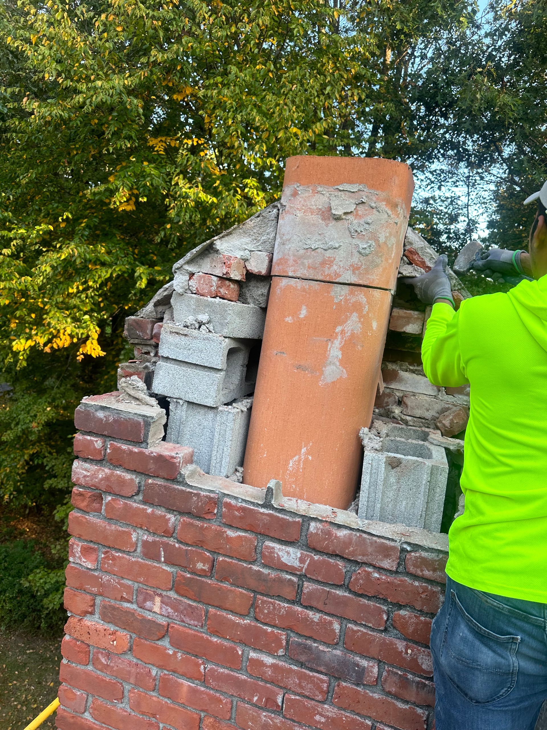 A worker in a neon yellow shirt repairs a brick chimney, exposing the interior flue and concrete block structure.