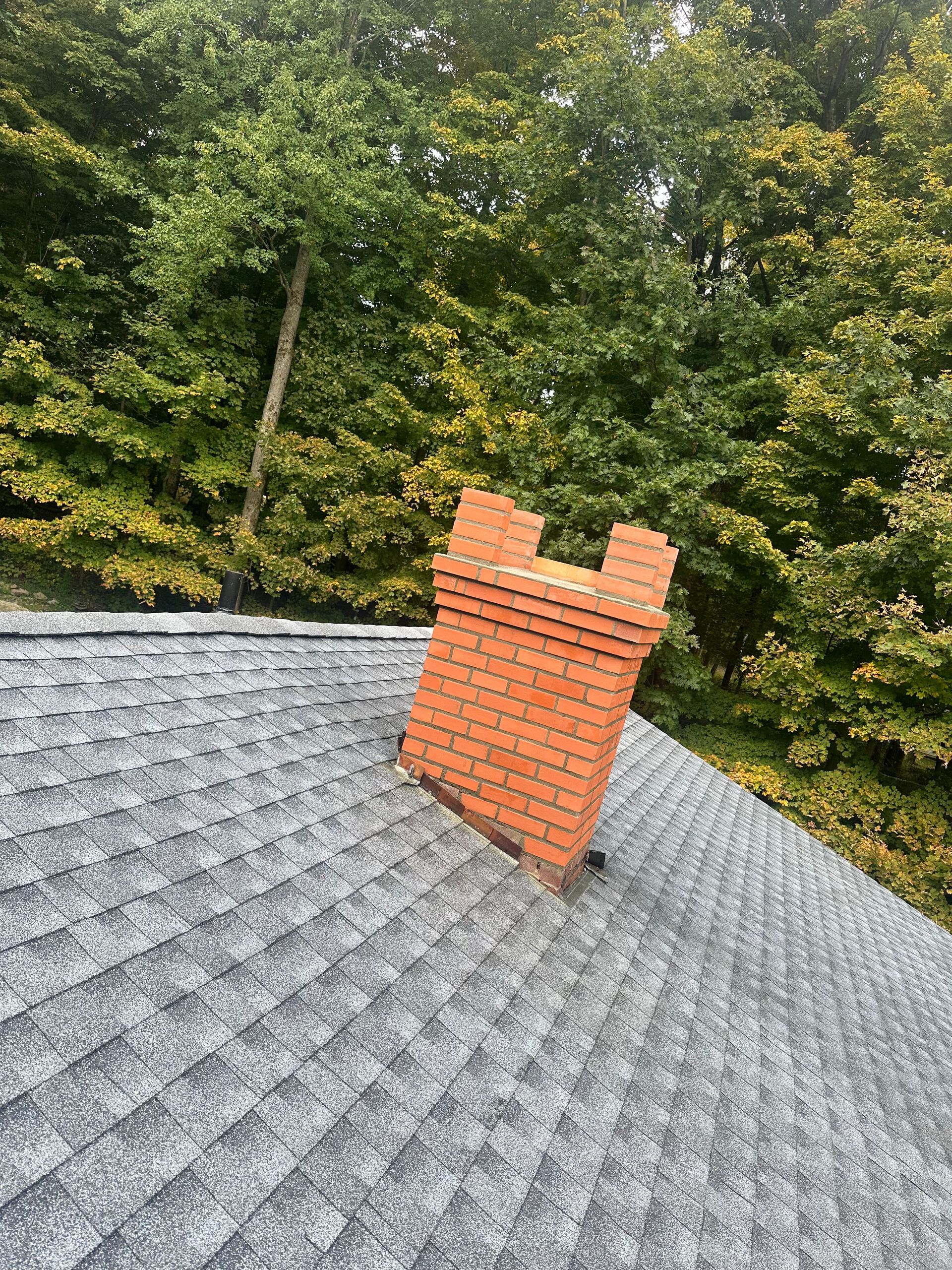 A red brick chimney rises from a gray asphalt shingled roof, set against a backdrop of green trees.