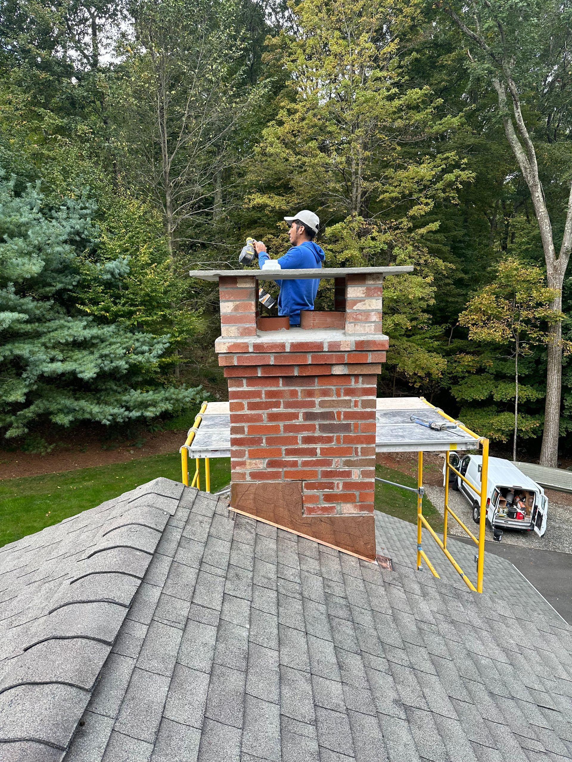A worker in a blue shirt repairs a red brick chimney on a house roof, supported by yellow scaffolding.