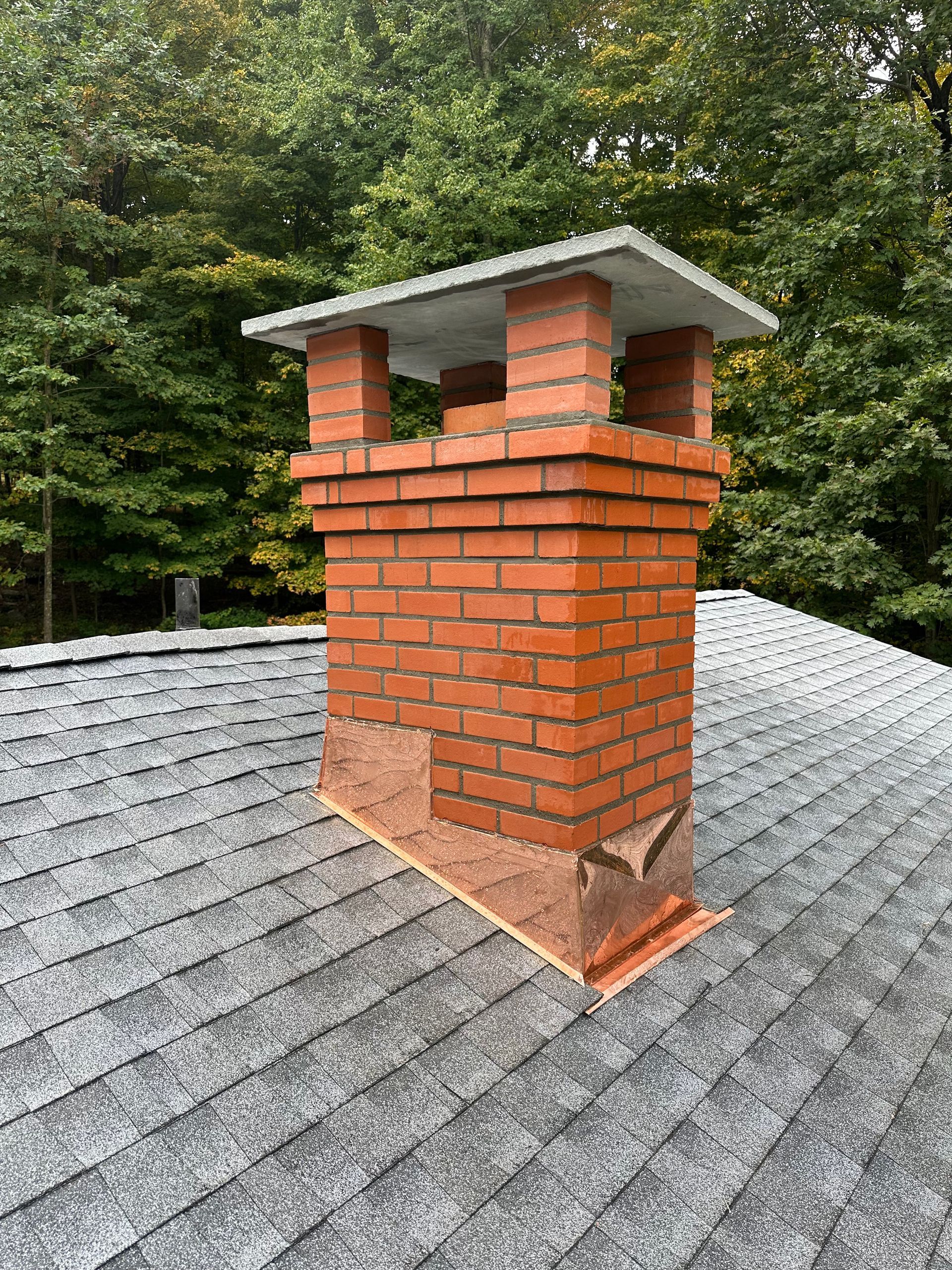 A red brick chimney with a stone cap sits on a grey asphalt shingled roof, set against a background of green trees.